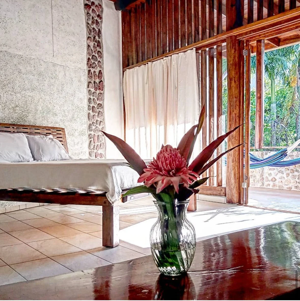 A cozy bedroom with a wooden bed, white bedding, and a flower arrangement in a glass vase on a wooden table. There's a veranda with wooden accents, open to a view of palm trees and a hammock outside.