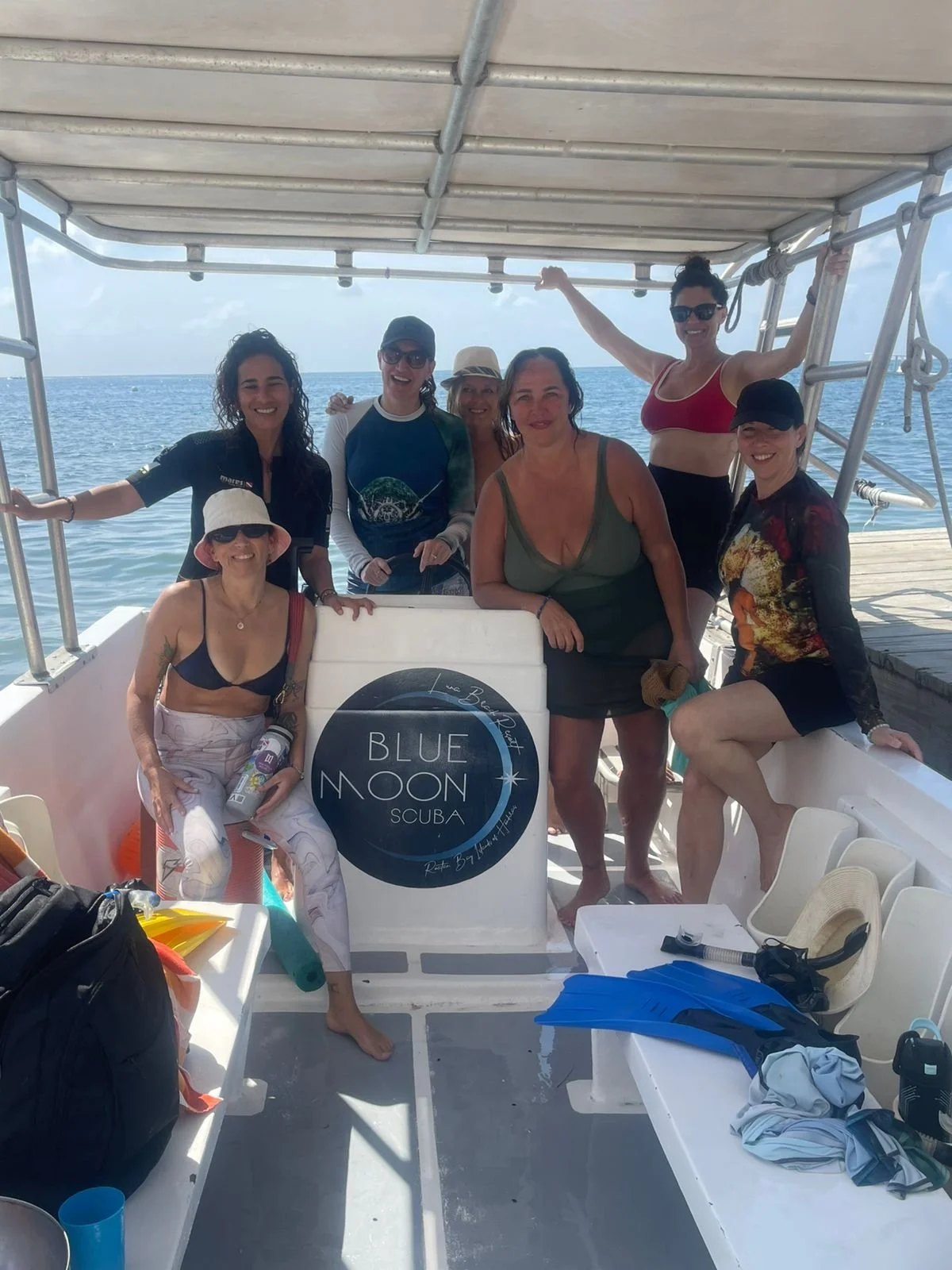 Group of women on a boat, posing with a sign that reads 'Blue Moon SCUBA', waterproof gear and snorkeling equipment visible, ocean in background.