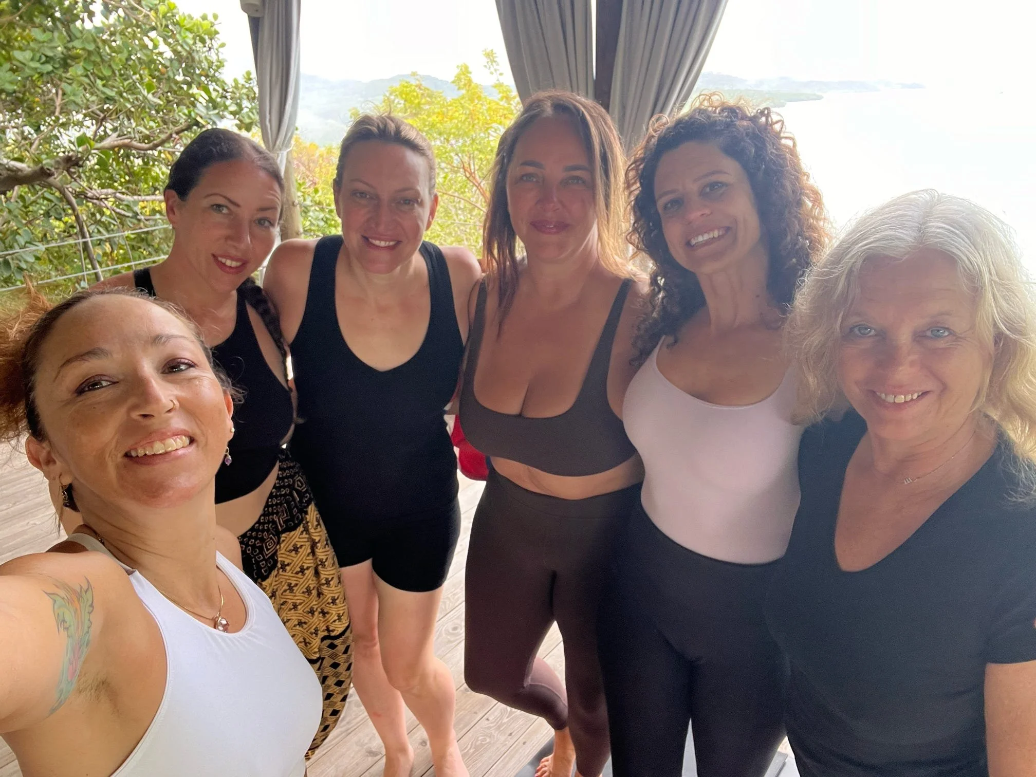 Group of seven women in fitness attire taking a selfie outdoors, with trees and cloudy sky in the background.