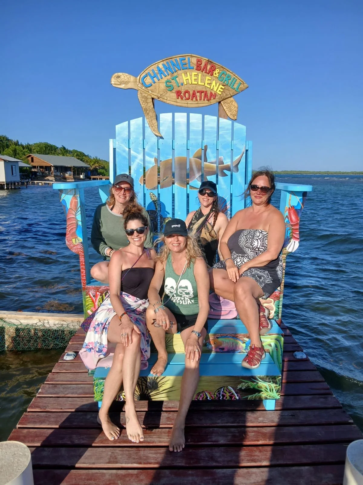 Five women sitting on a colorful bench with a nautical theme, by the water, with huts and a blue sky in the background.