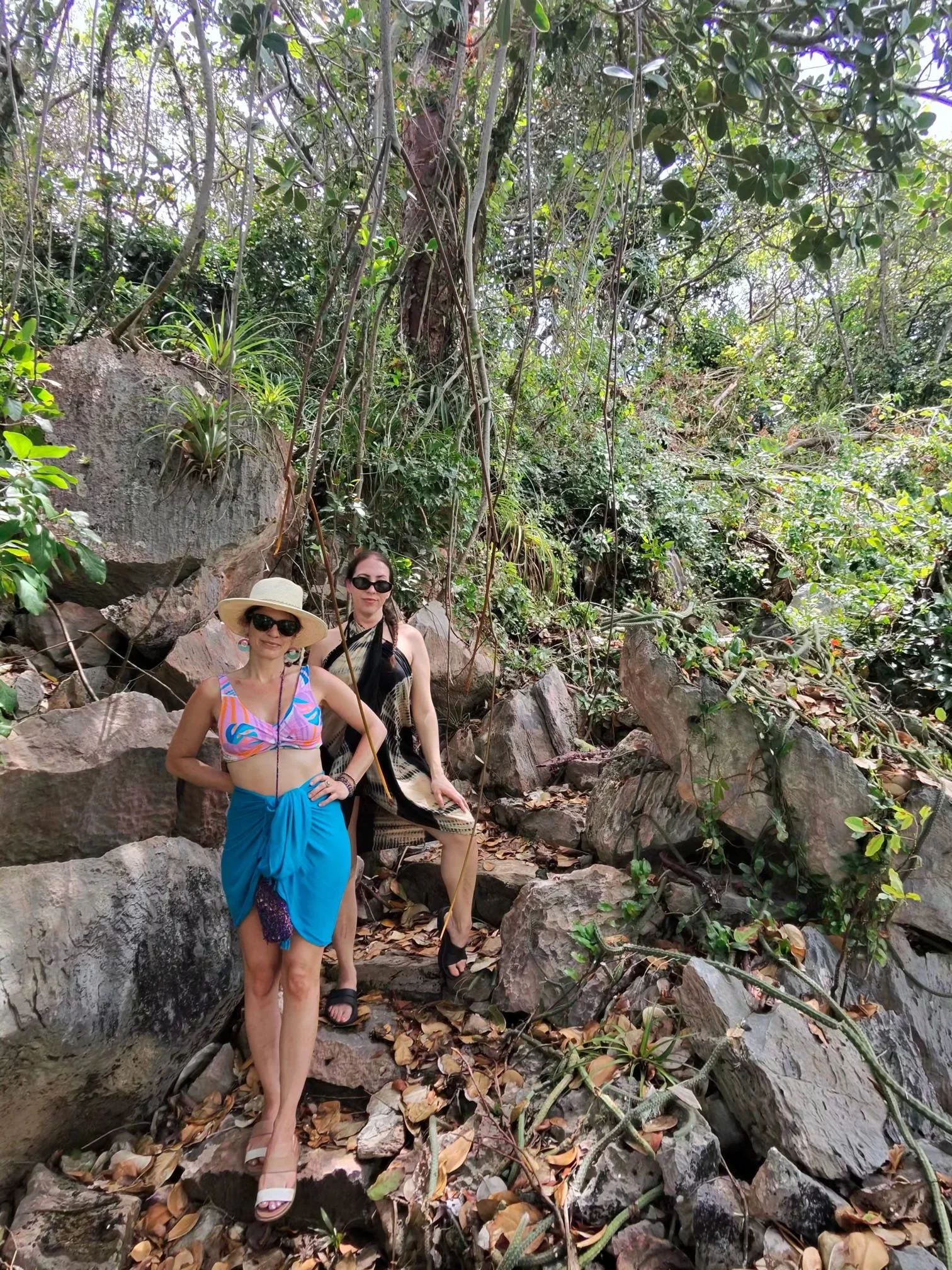 Two women standing on rocky terrain surrounded by lush greenery in a forest or jungle setting.