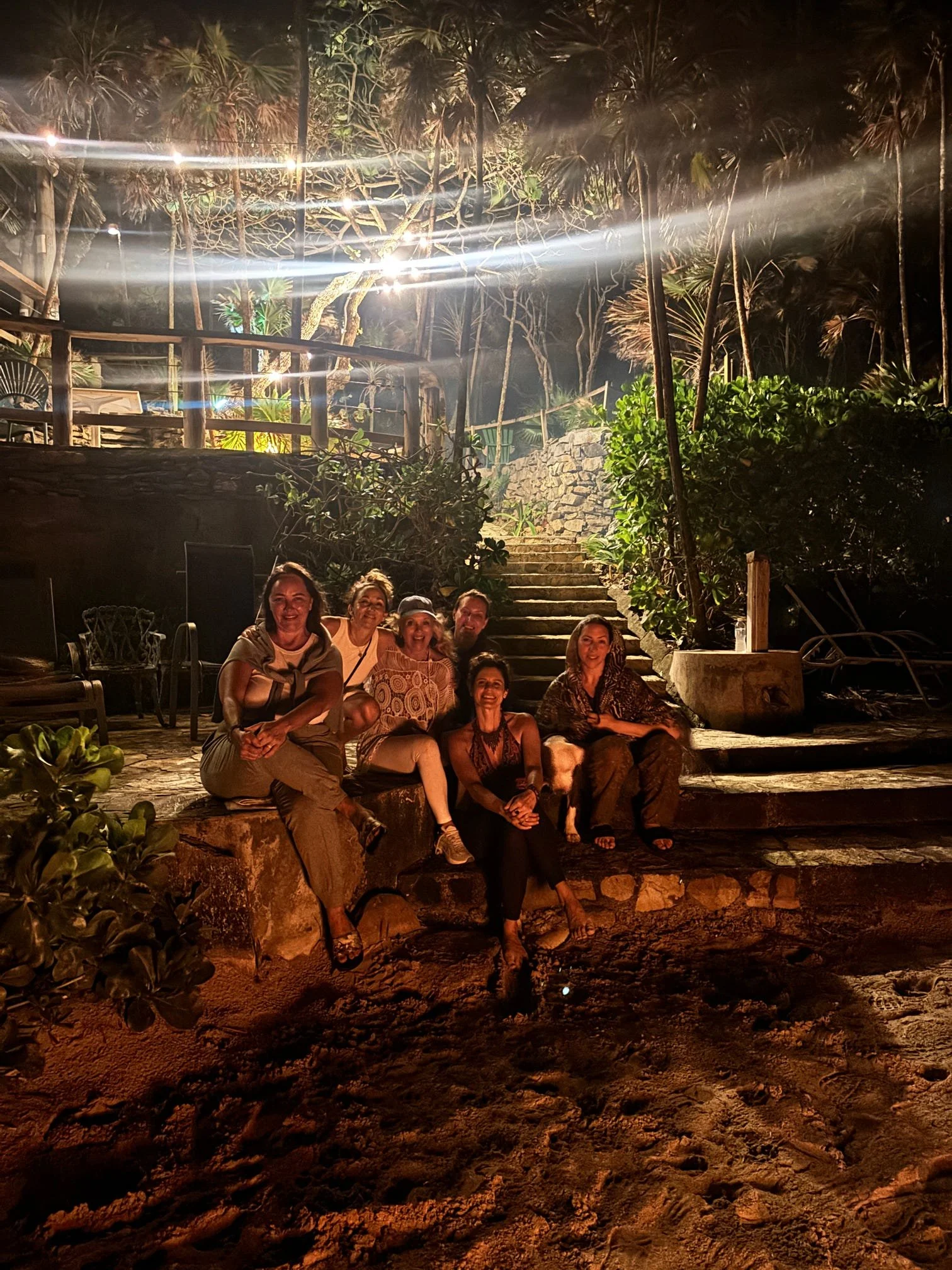Group of six people sitting on rocks and steps outdoors at night, illuminated by bright lights, with trees and greenery in the background.