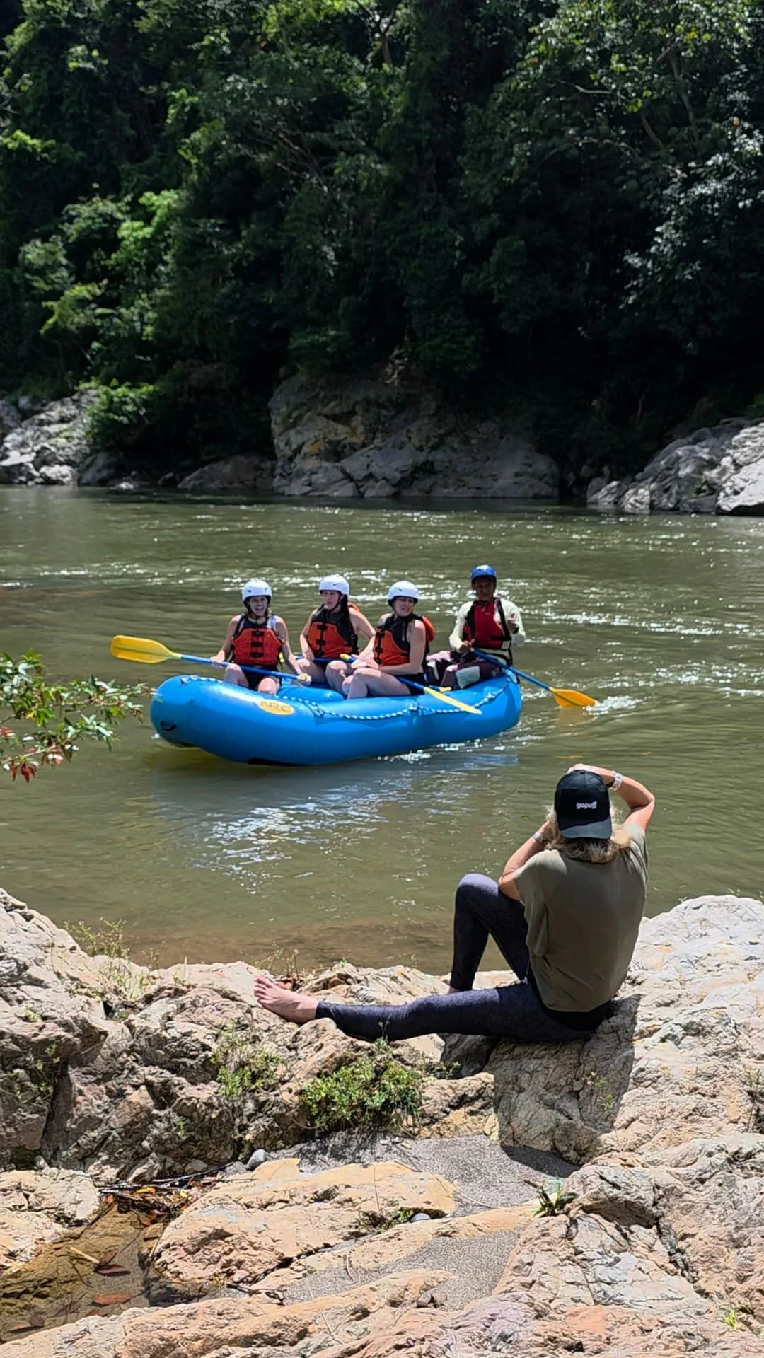 Four people in a blue inflatable raft with life jackets and helmets, paddling on a river surrounded by trees and rocks. A person sitting on the rocky shore takes a photo or videos of the scene.
