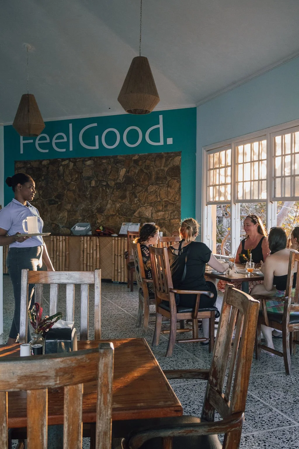 Interior of a restaurant with a large window letting in natural light, wooden tables and chairs, a stone accent wall, and a sign on the wall that says "Feel Good." Four women are seated at a table, enjoying a conversation, while a waitress in a white
