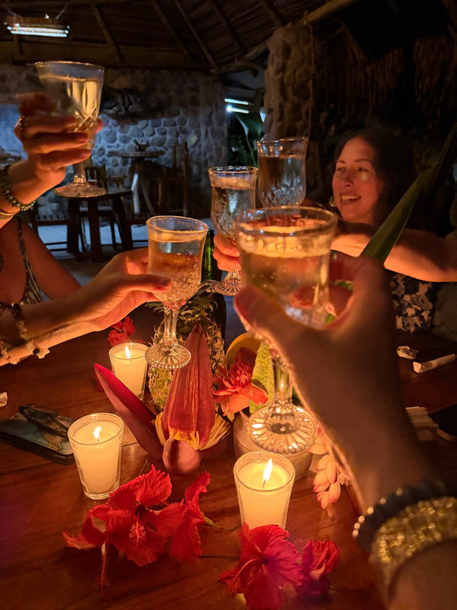 Friends celebrating with champagne glasses raised in a toast around a decorated table with candles, flowers, and tropical decor in a cozy, rustic setting.