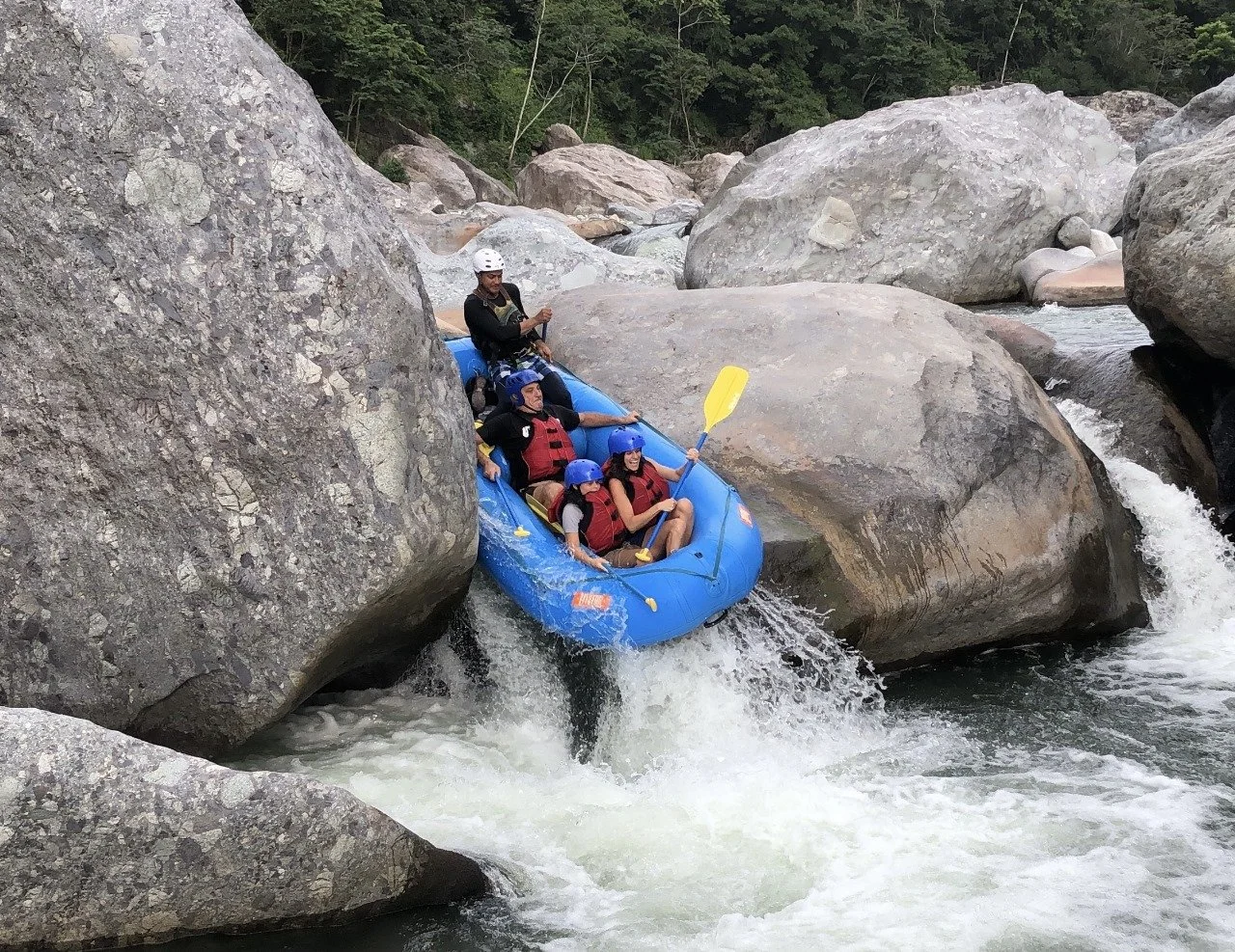 Four people in a blue inflatable raft wearing helmets and life vests navigating through a rocky river with large boulders.