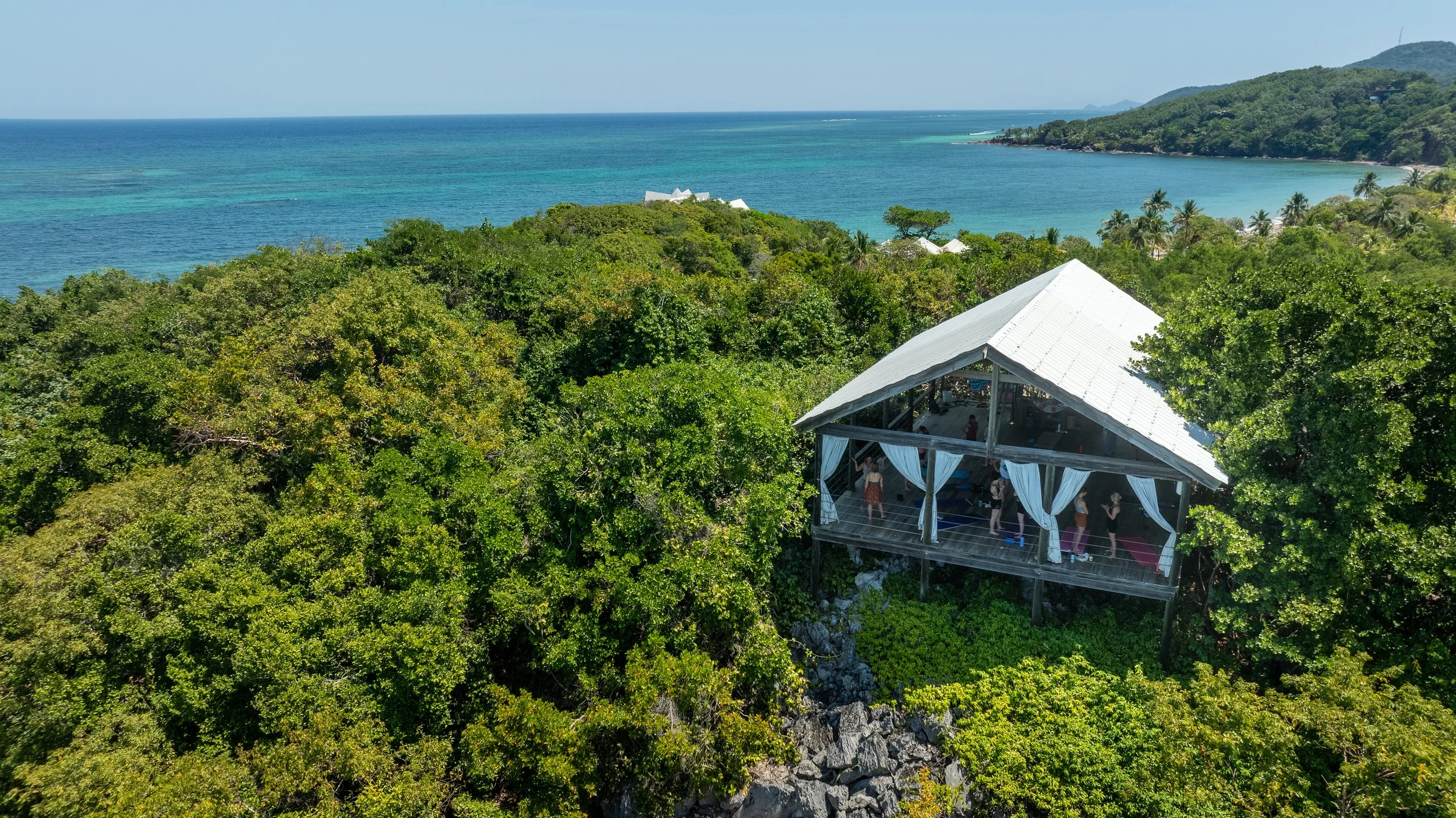 A wooden structure with curtains on a hillside overlooking the ocean, surrounded by lush green trees and a distant view of a coastal landscape.