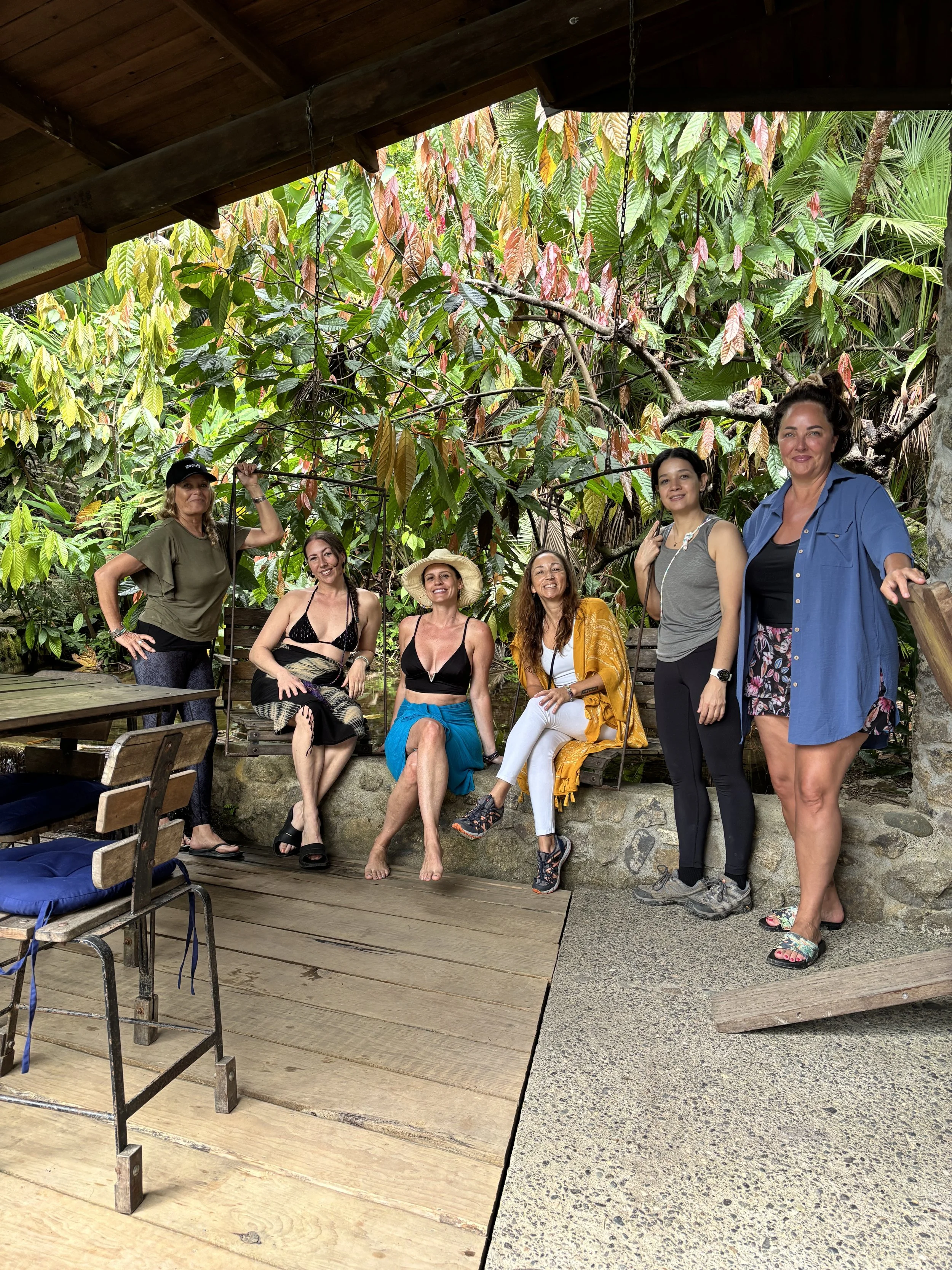 Six women posing together outdoors near lush tropical plants, some sitting on a wooden swing, others standing, in a garden setting.