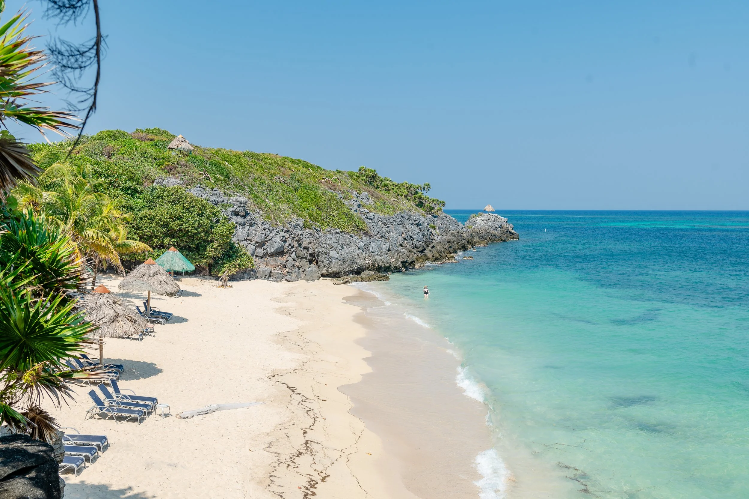 Tropical beach with white sand, lush green vegetation, thatched umbrellas, lounge chairs, and clear turquoise water with a rocky hillside in the background.