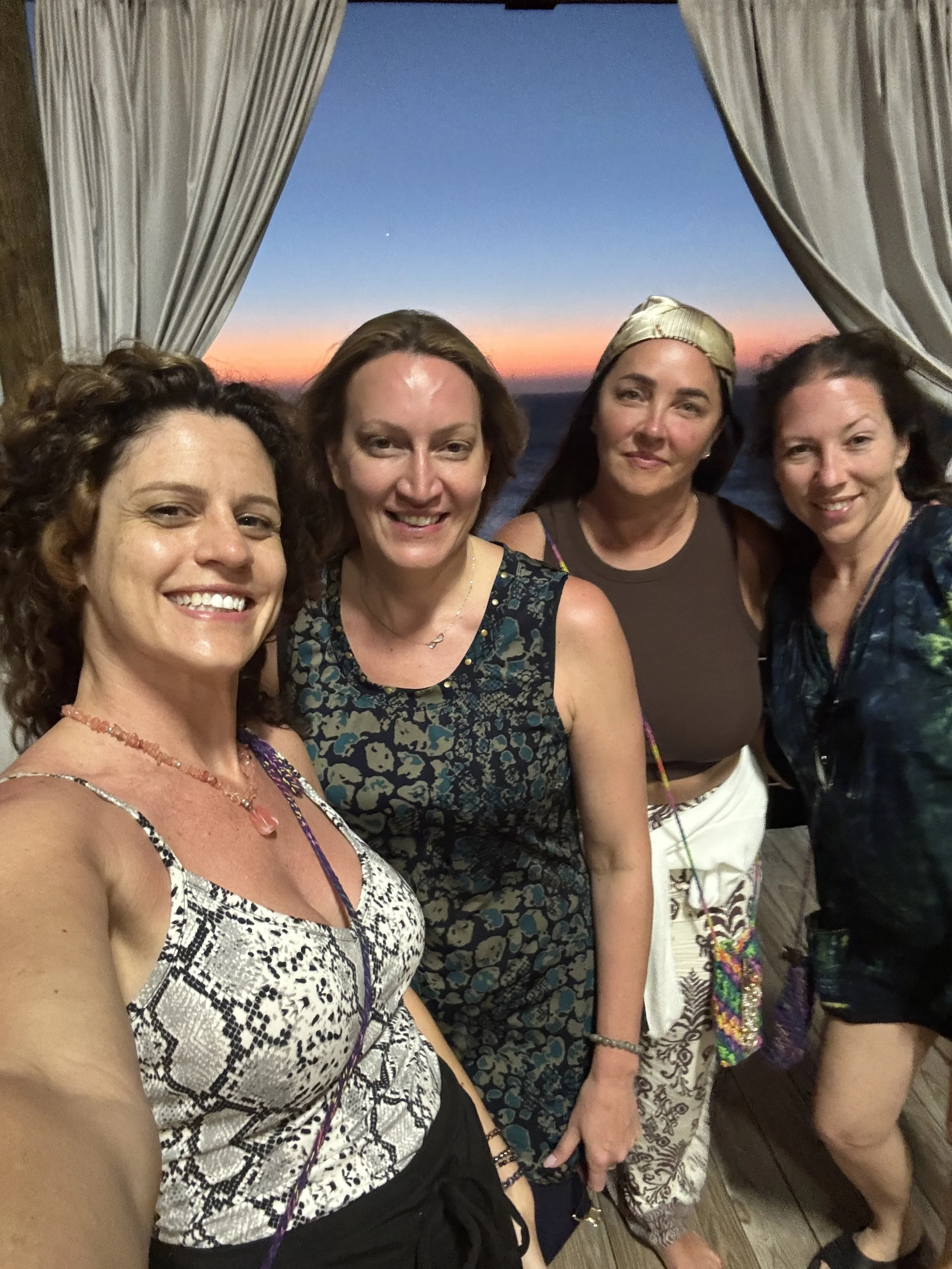 Four women with curly and straight hair taking a selfie together at sunset, with curtains and the ocean in the background.