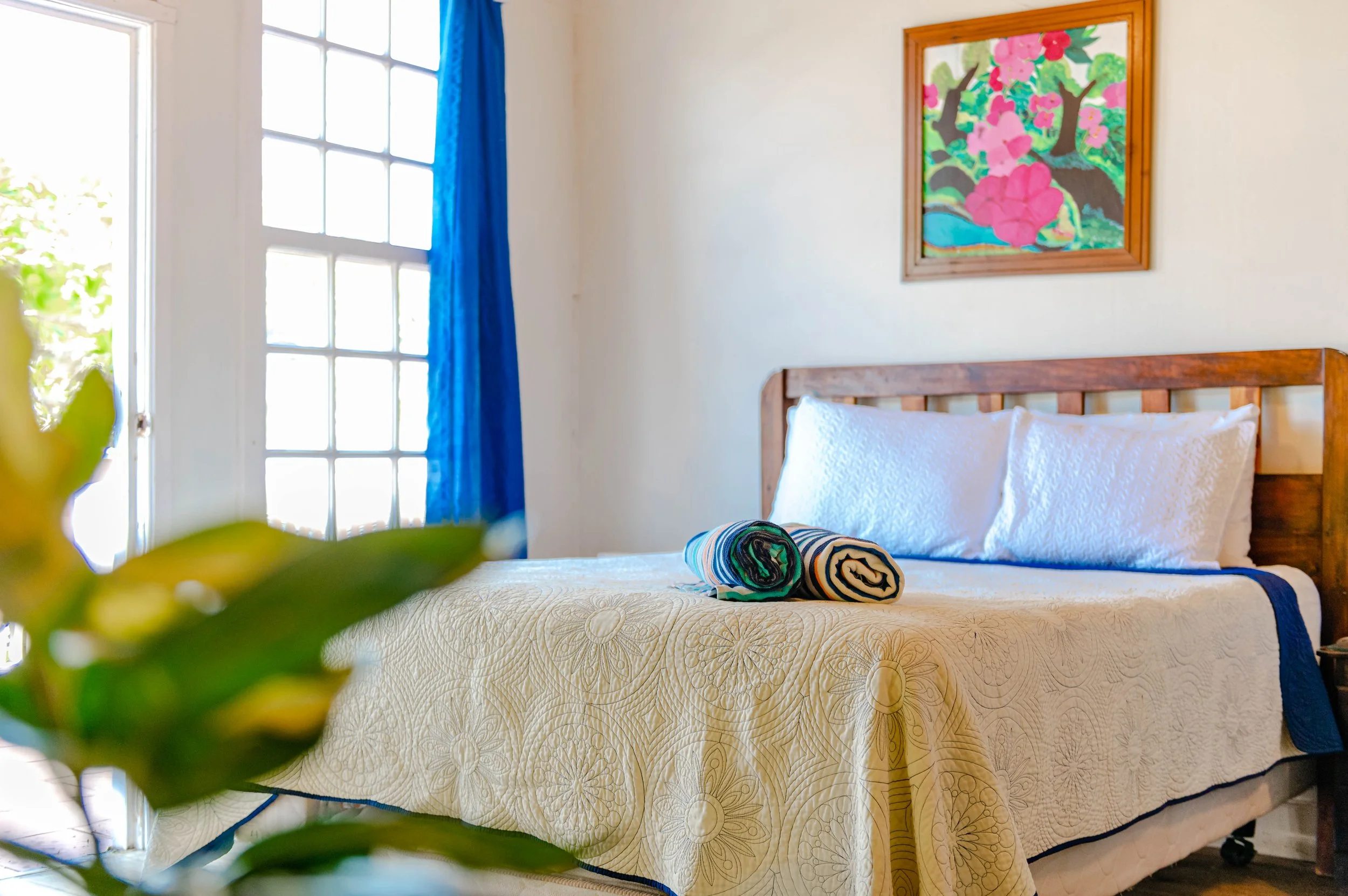 Clean hotel room with a bed covered in a cream quilt, two rolled towels on top, and a wooden headboard. A colorful painting of pink flowers and green trees hangs above the bed. There is a large window with blue curtains to the left, allowing natural