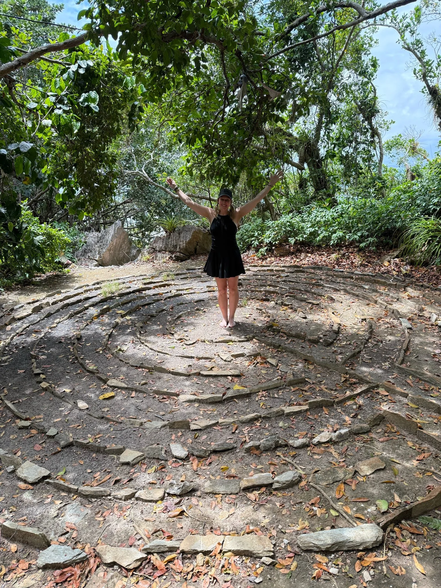 A woman in a black dress and cap standing with arms raised on a spiral stone path in a lush forest.