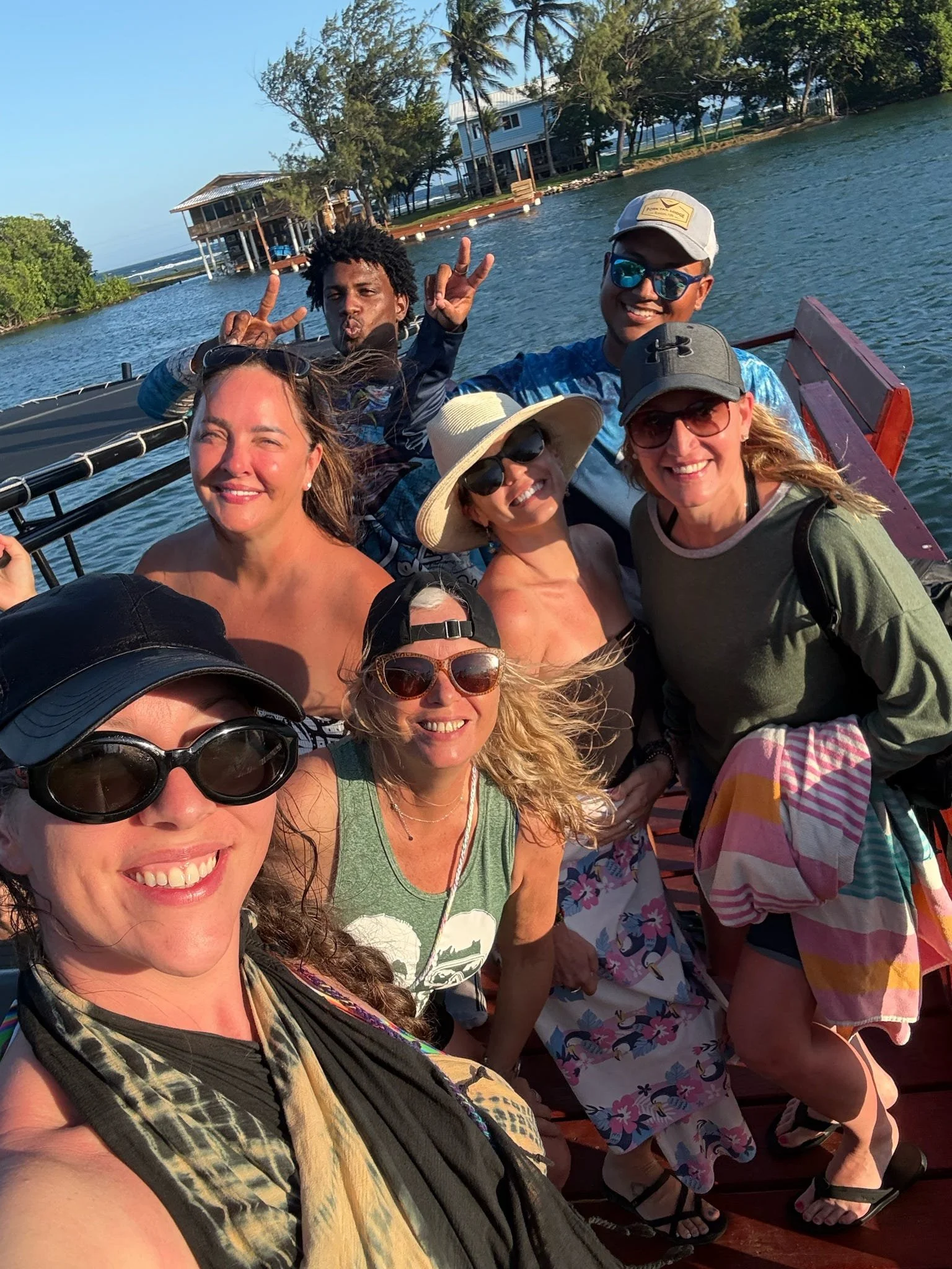 Group of seven friends on a boat taking a selfie with water and trees in the background, smiling and wearing summer clothes and hats.