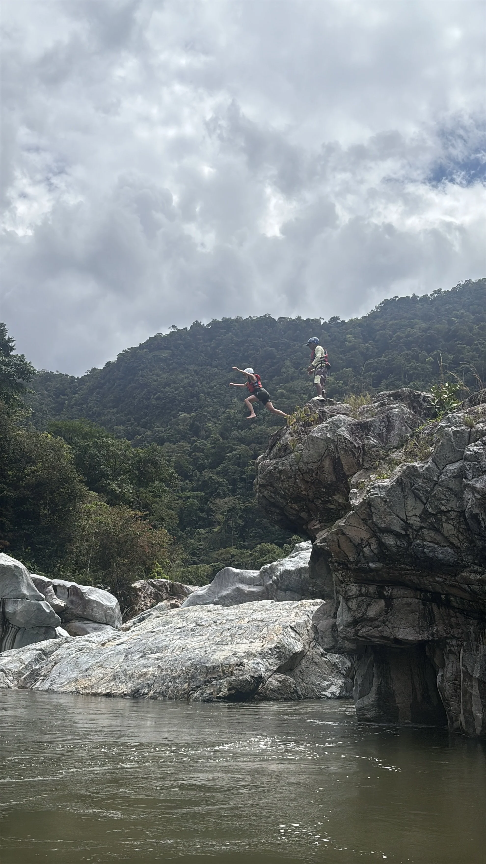 Two children jump off a large rock into a river surrounded by mountains and trees, with a cloudy sky overhead.