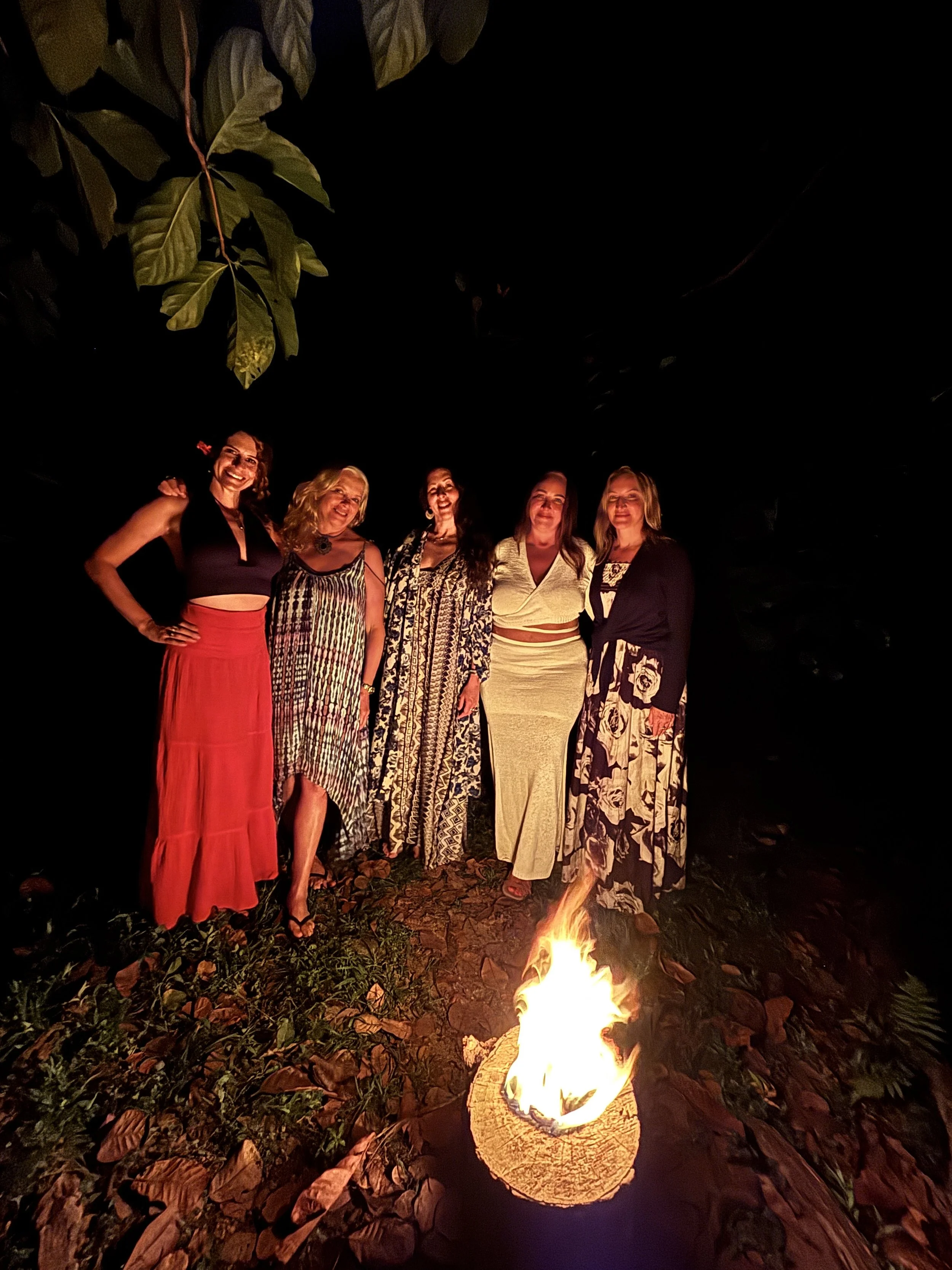 Five women standing outdoors at night near a small fire, smiling, surrounded by dark foliage.