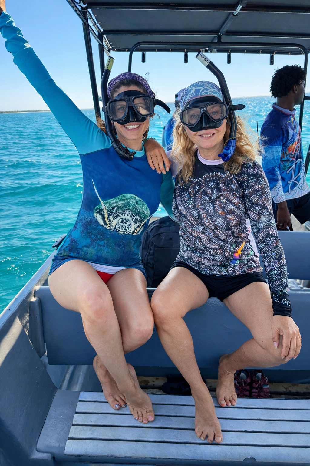 Two women sitting on a boat, wearing snorkeling gear and swimwear, smiling at the camera against a blue ocean background.