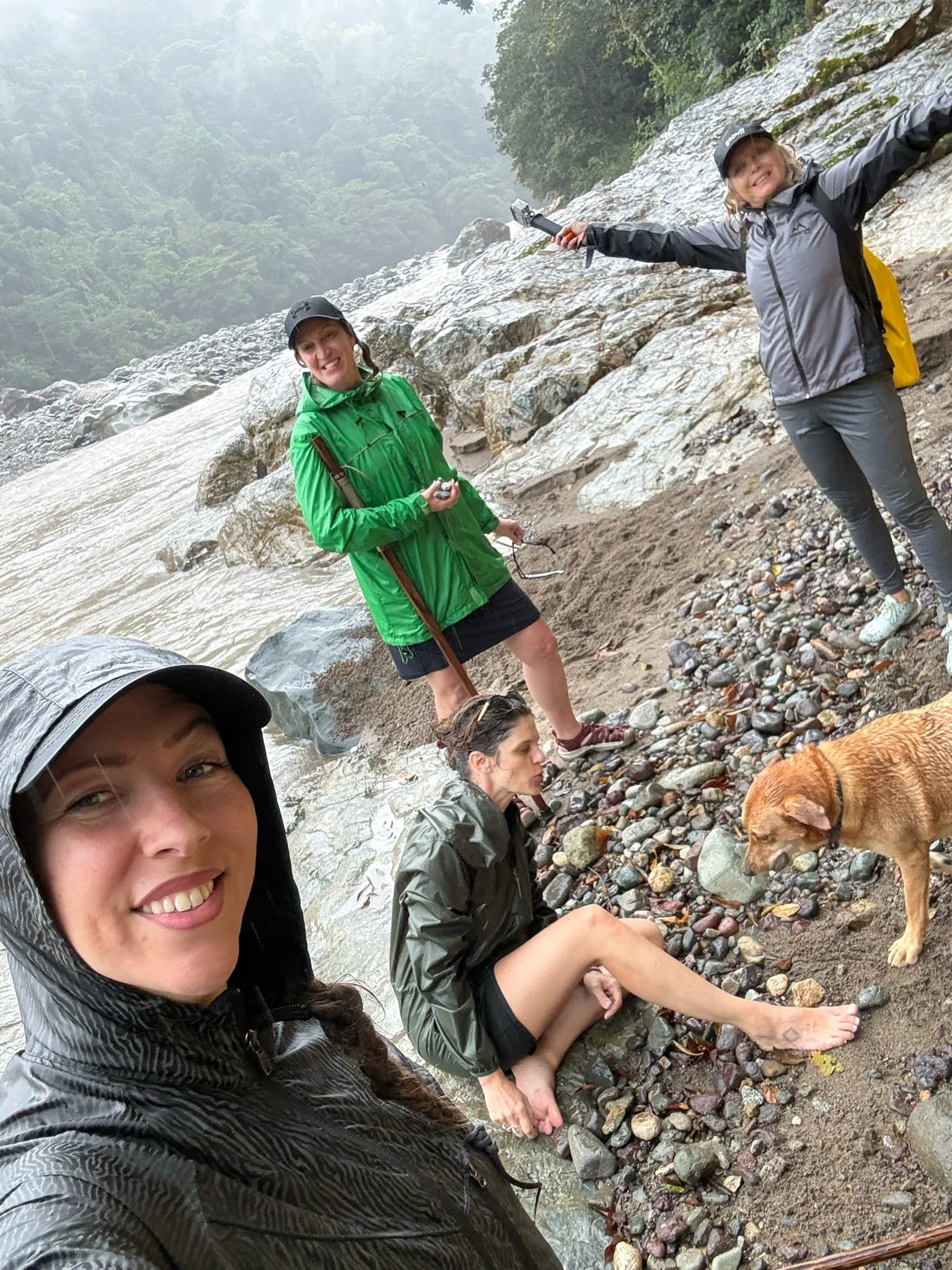 Four women and a dog on a rocky and muddy riverbank in a misty forested area, some wearing rain jackets and hats, with one woman taking a selfie in the foreground.