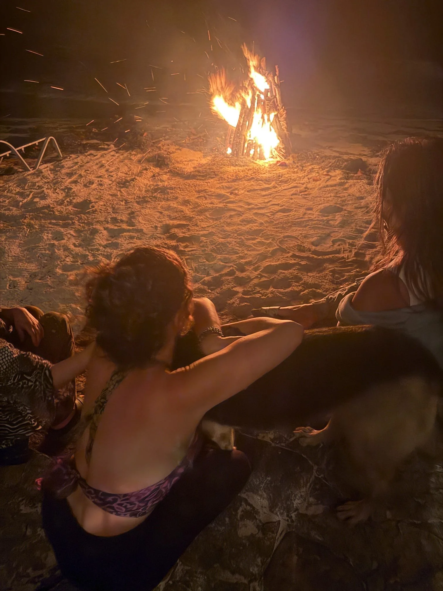 People sitting on the beach watching a bonfire at night.