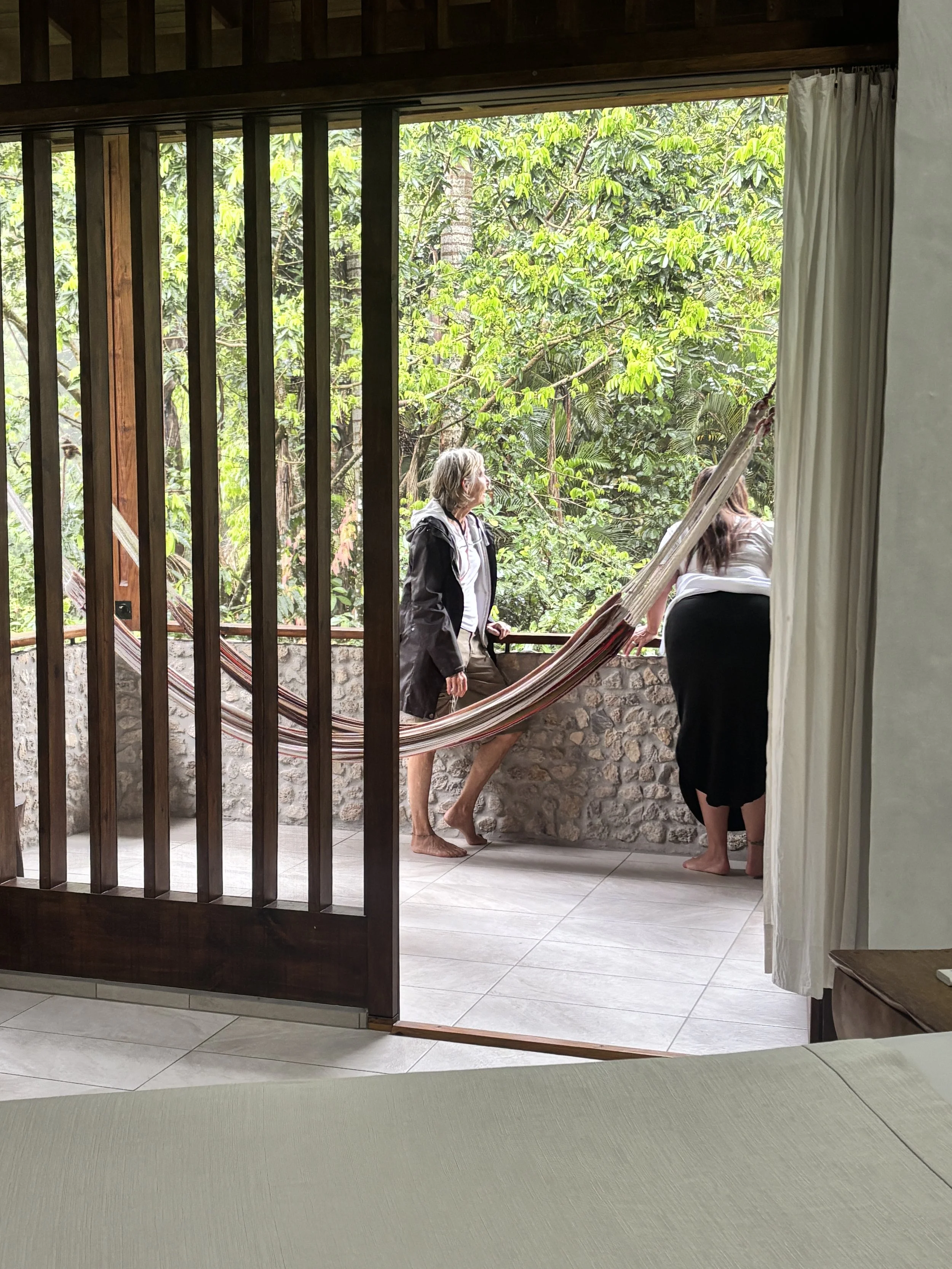 Two women and one man standing on a balcony with a hammock, surrounded by green trees, seen from inside a room.
