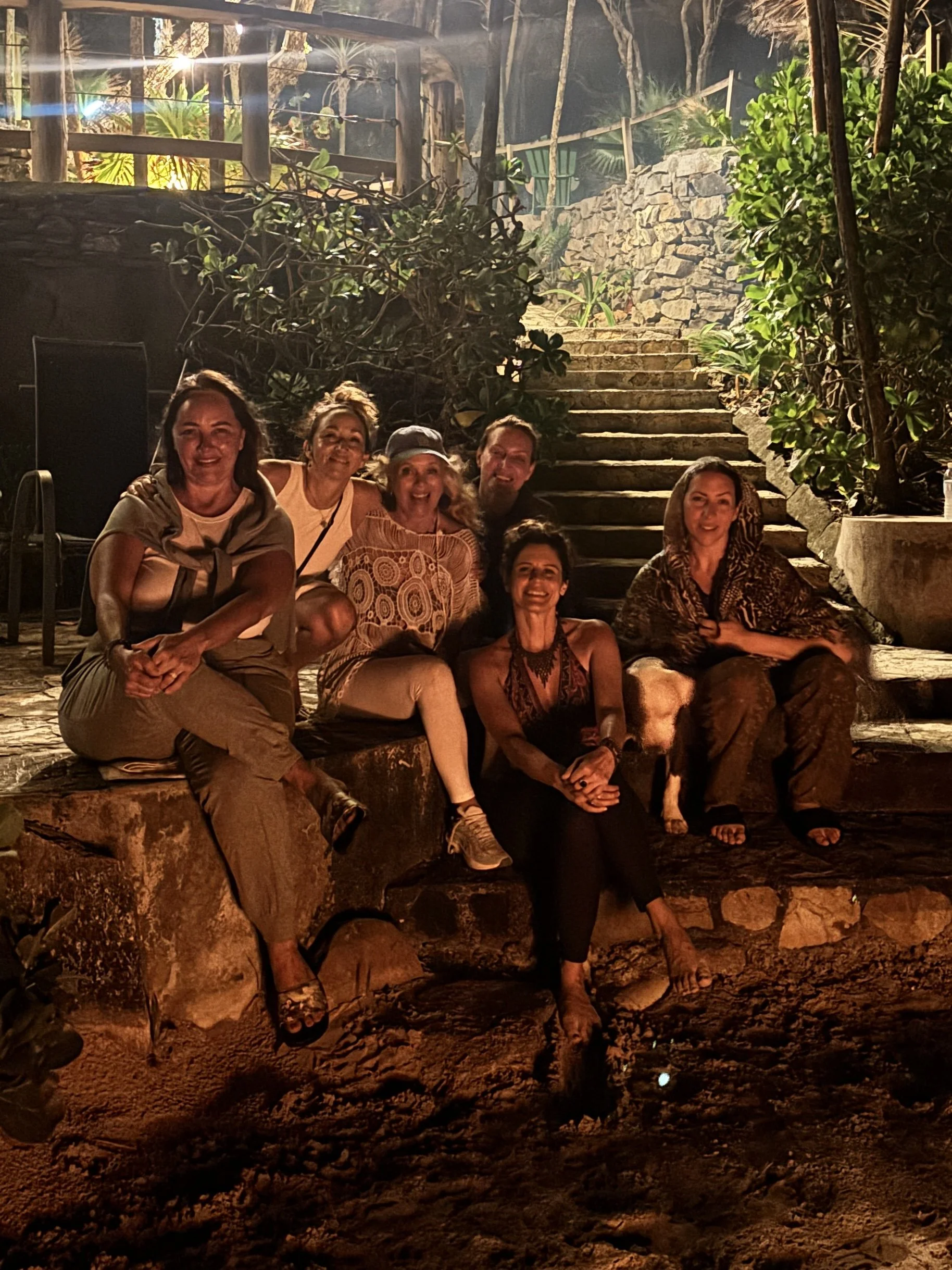 Group of six women and one man sitting on stone steps outdoors at night, illuminated by warm lighting, surrounded by lush greenery and rocks.