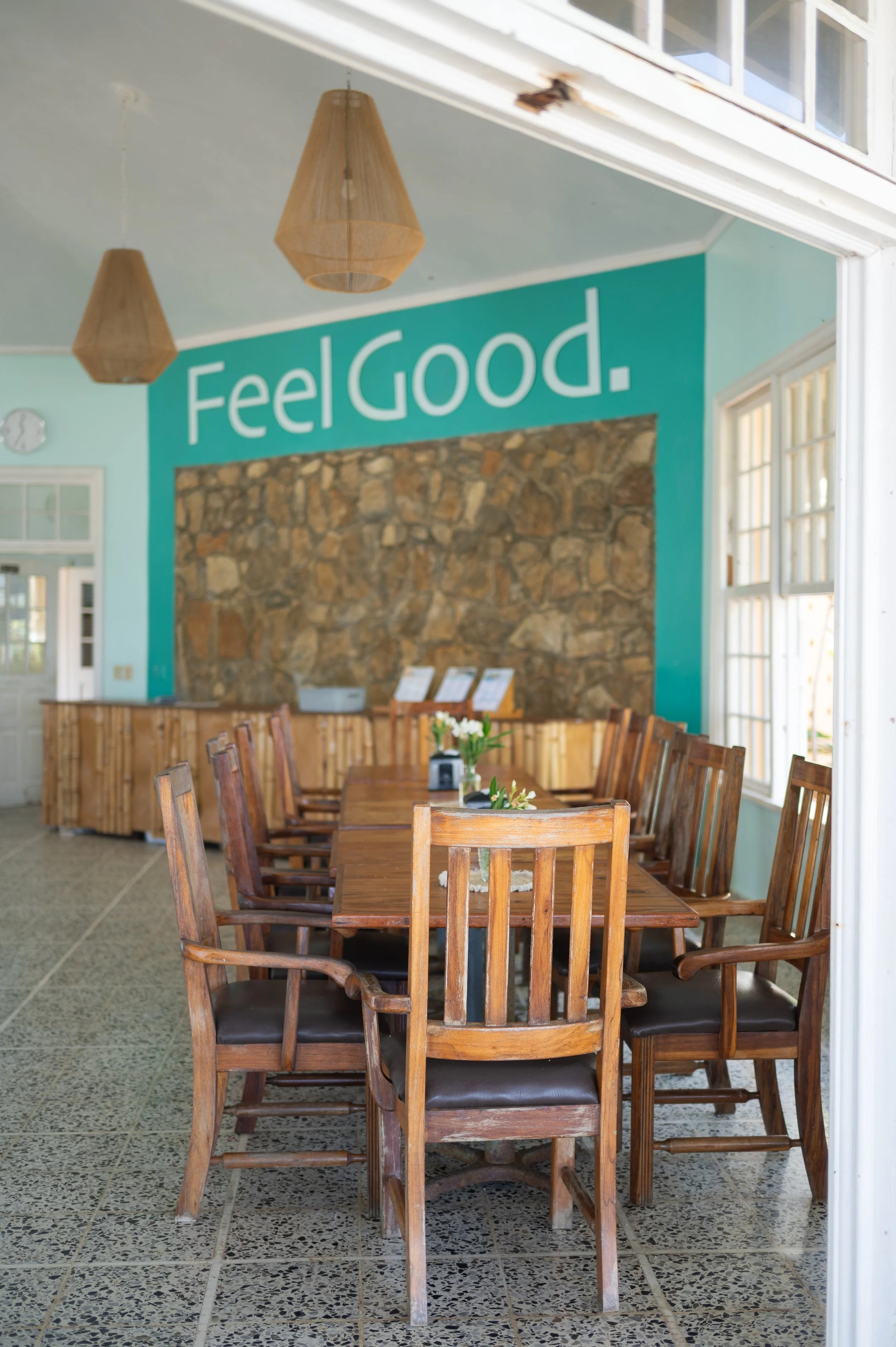 Interior of a dining room with a long wooden table and multiple wooden chairs with black cushions, a turquoise wall with the words 'Feel Good' written on it, and a stone accent wall behind the table.