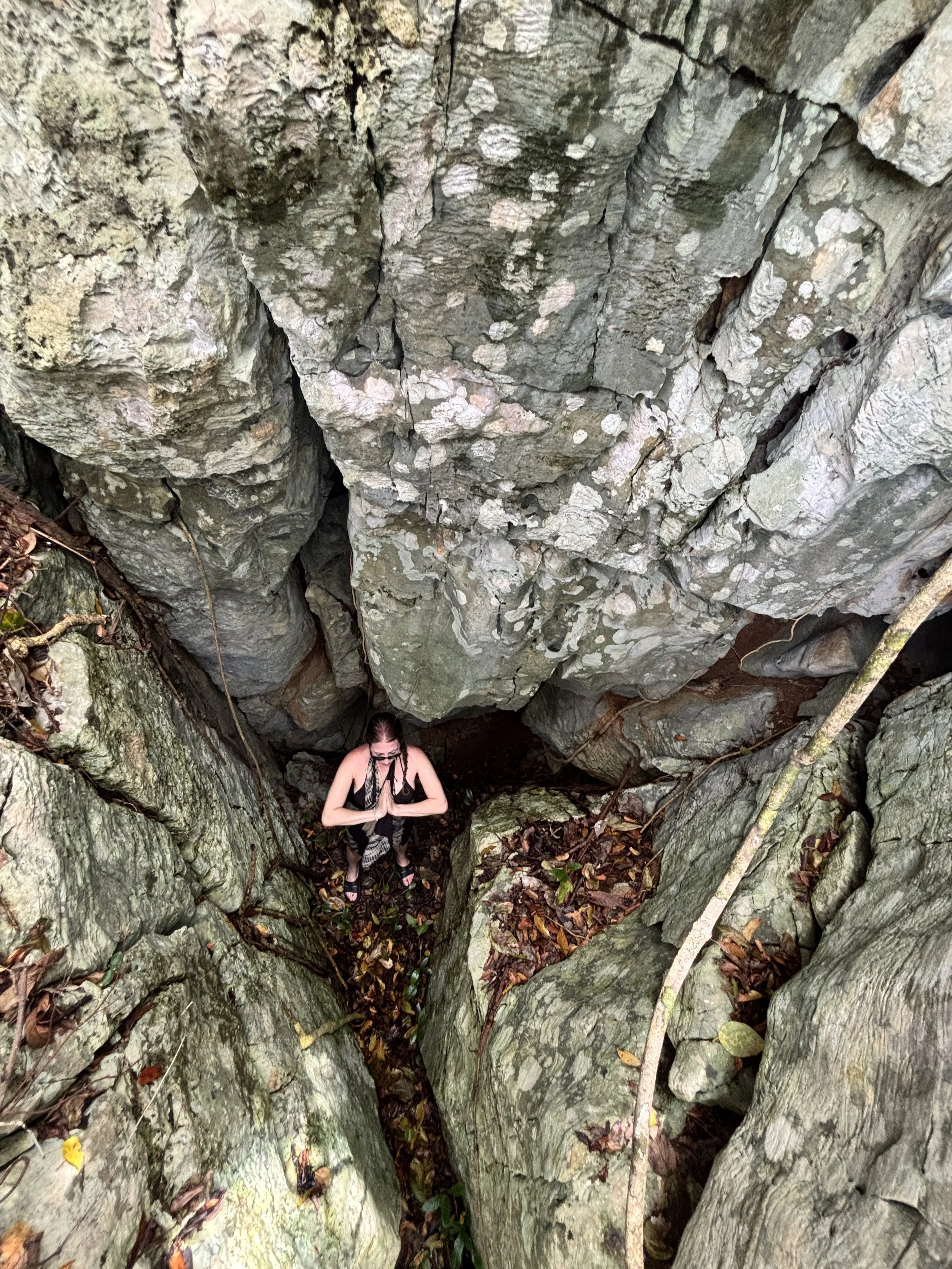 A person with braided hair and glasses standing in a rocky crevice with large gray boulders and dried leaves around them, hands pressed together in a praying gesture.