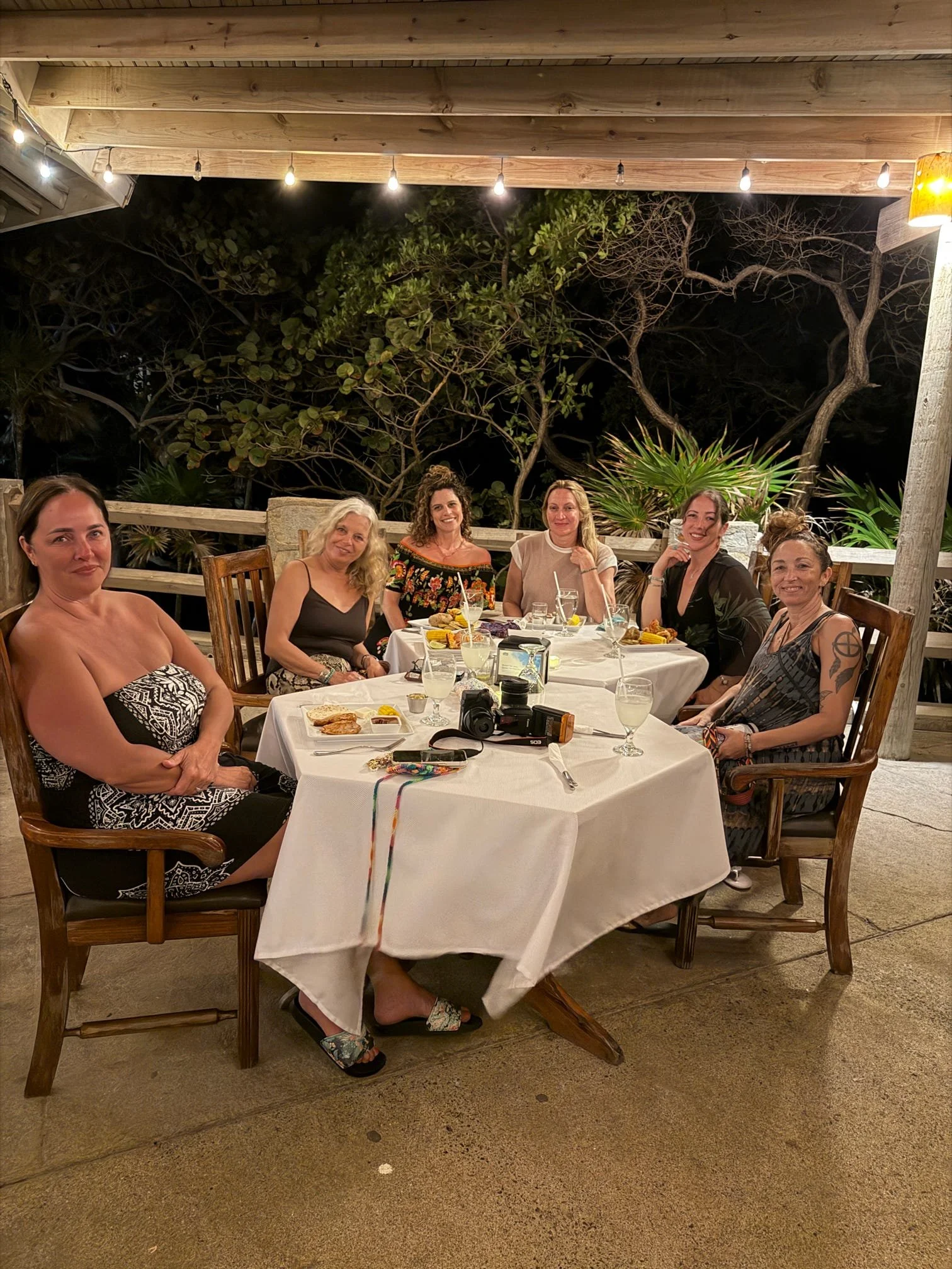 Group of six women sitting around a table at an outdoor restaurant at night, with some food and drinks on the table, and trees in the background.