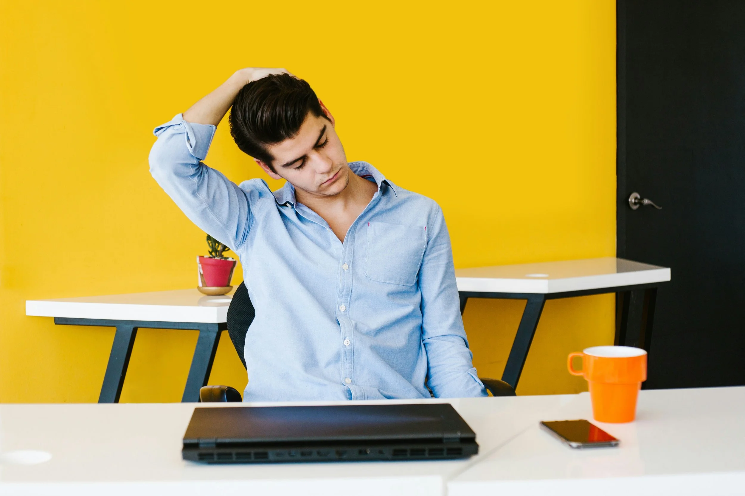 Young man sitting at a desk with a closed laptop, looking down with a pained expression, hand on his head, in a brightly colored office with yellow wall and a small plant in a pink pot.