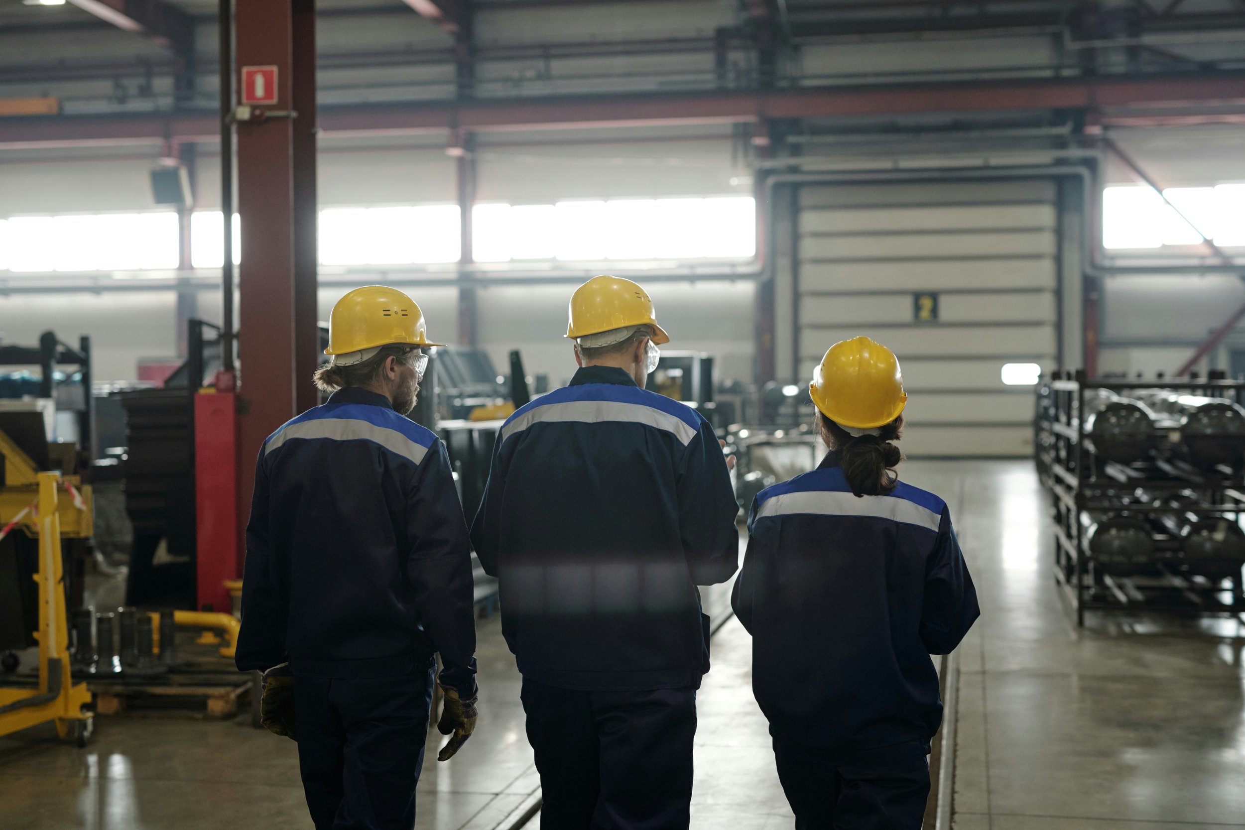 Three factory workers wearing yellow safety helmets and blue uniforms walking through an industrial warehouse.