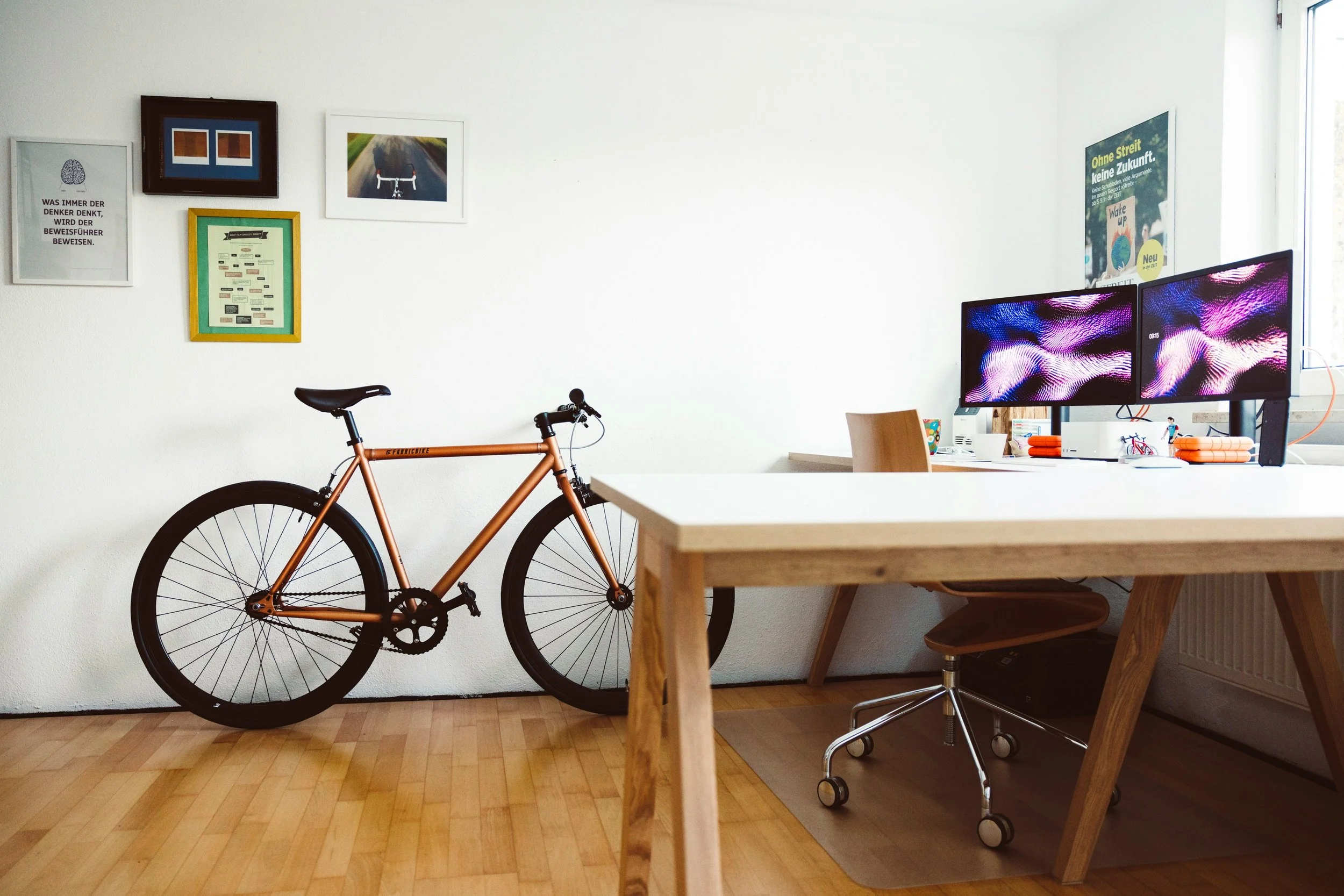A room with a peach-colored bicycle leaning against a white wall, a wooden desk with dual monitors, a wooden chair, and several framed pictures and posters on the wall.