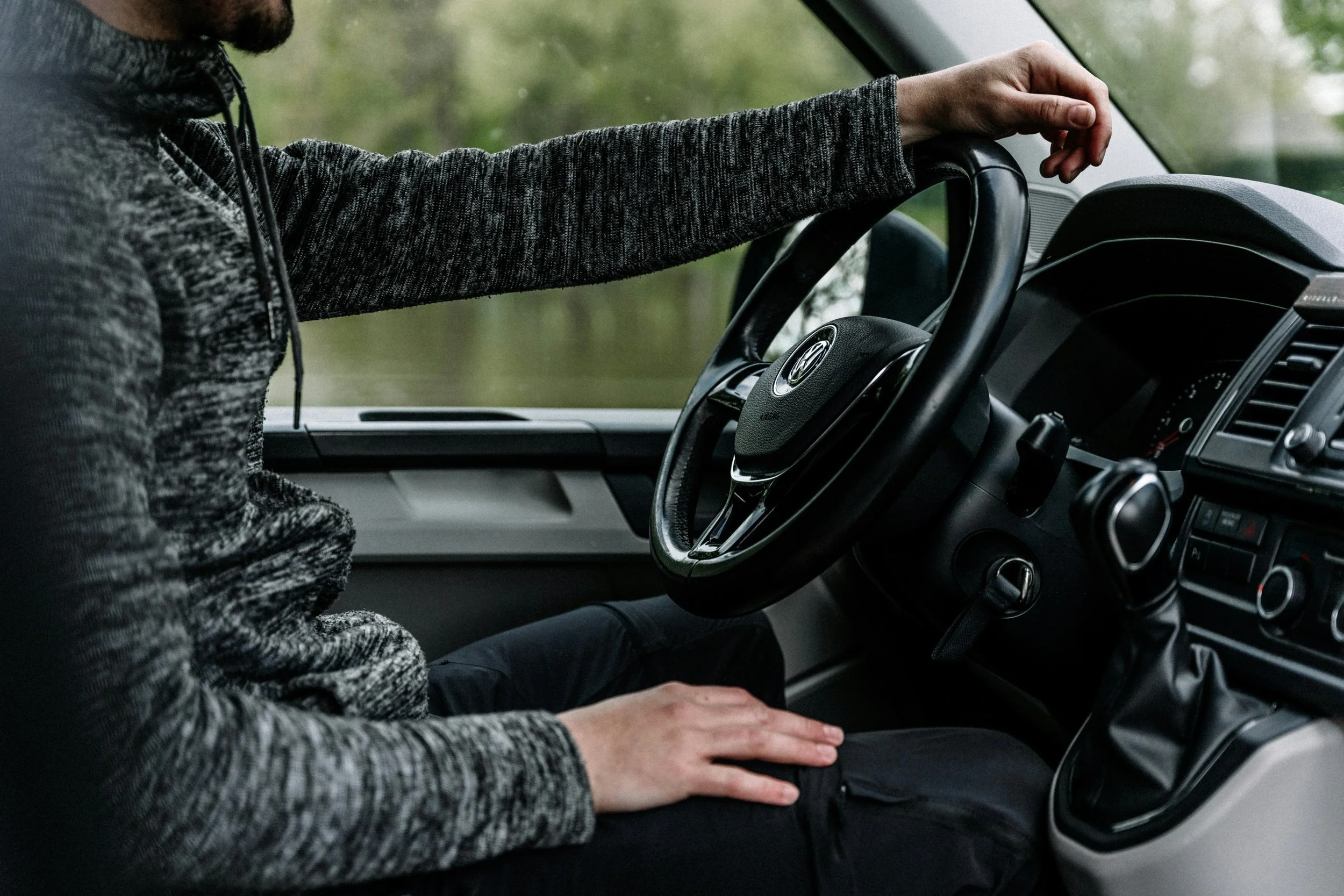 Person wearing a gray sweater driving a van, with their left hand on the steering wheel and right hand resting on their lap. The dashboard and gear shift of the van are visible.