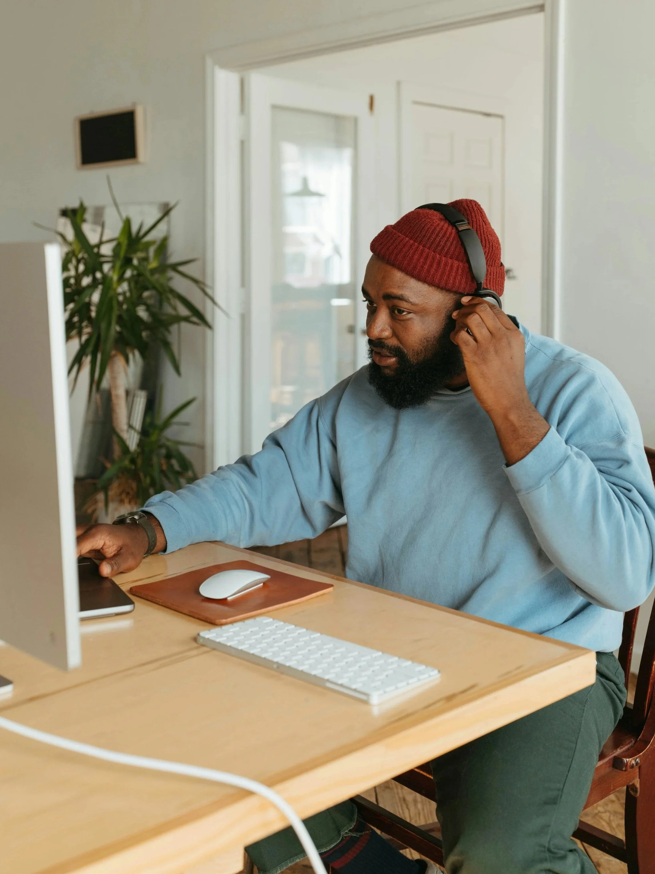 A man with a beard, wearing a red beanie and blue sweatshirt, wearing headphones, working on a computer at a wooden desk in a room with plants and a door with glass panels.