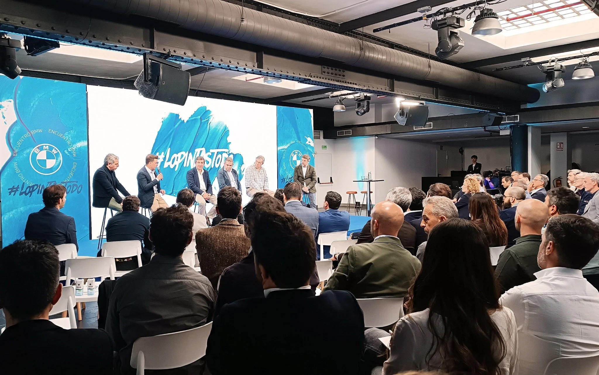 Un panel de seis personas hablando en un escenario durante una conferencia, con una audiencia sentada en sillas blancas, en un ambiente moderno con pantallas grandes y equipo técnico visible.