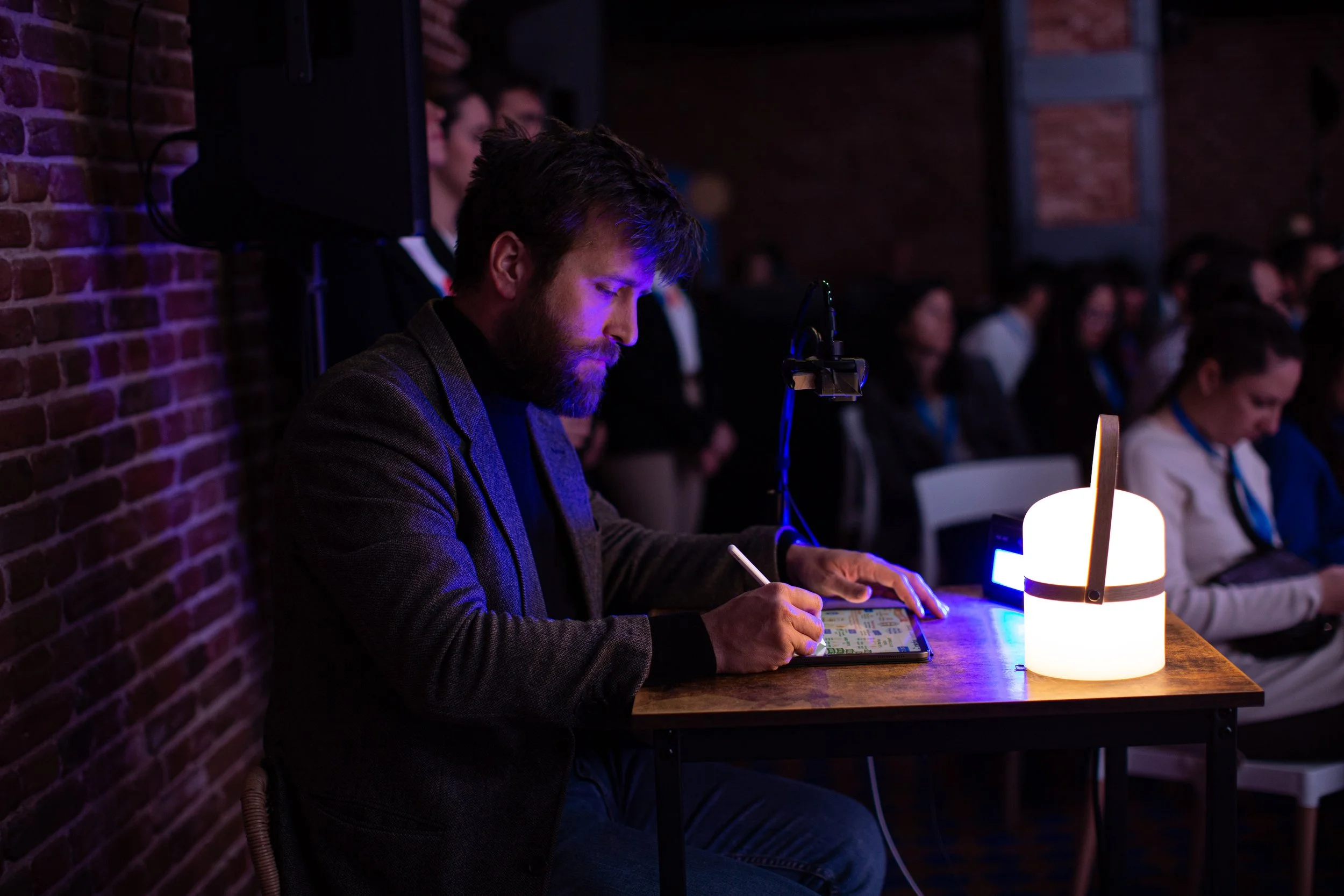 Hombre con barba sentado en una mesa de madera en un lugar oscuro con iluminación tenue, usando un bolígrafo y una tableta digital, con una lámpara moderna encendida frente a él y personas sentadas en el fondo en un entorno de evento o conferencia.