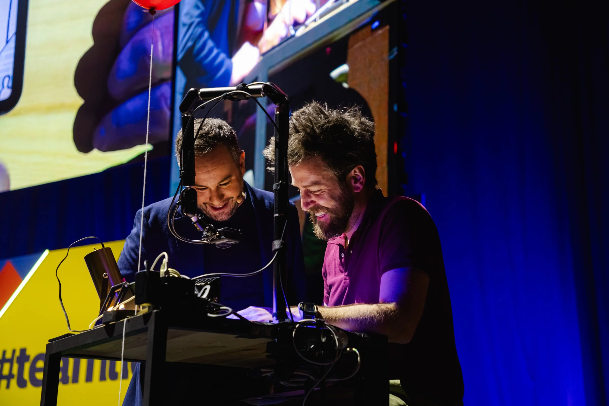 Javi Herrero (Grillo Visual Thinking) y el ilusionista mago Jorge Blass en escenario riendo y compartiendo una experiencia, con equipo de tecnología en una mesa, y una pantalla grande detrás mostrando una de las personas en la escena.