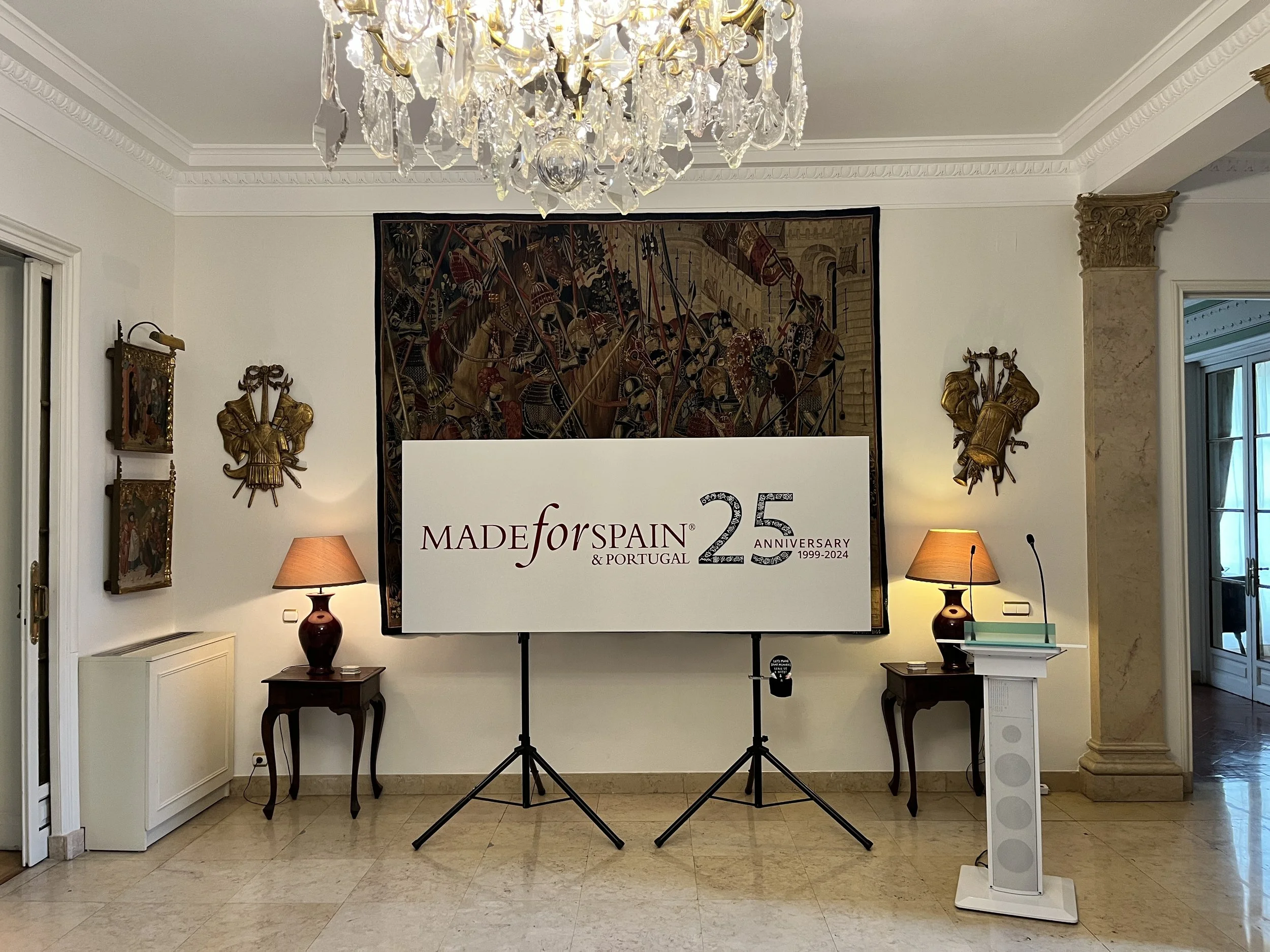 Sala elegante con un candelabro de cristal en el techo, dos mesas con lámparas y libros, un marco grande con arte en la pared trasera y un cartel que celebra los 25 años de MADE FOR SPAIN & PORTUGAL, 1999-2024