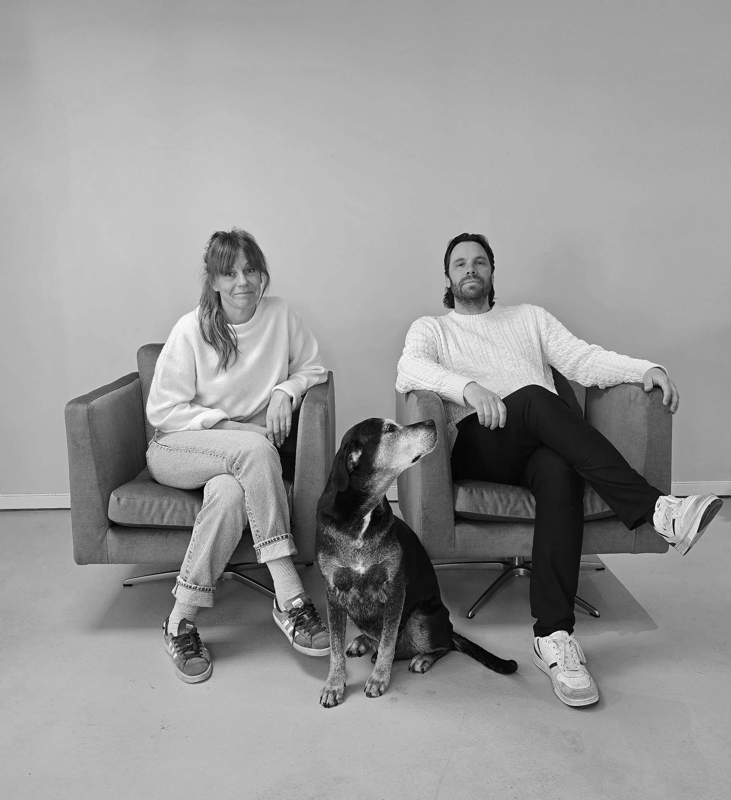 Två personer sitter i fåtöljer med en hund mellan sig. Kvinna till vänster och man till höger, alla i vita tröjor. Hundens huvud är inte synligt, men den sitter med kroppen vänd mot kameran.