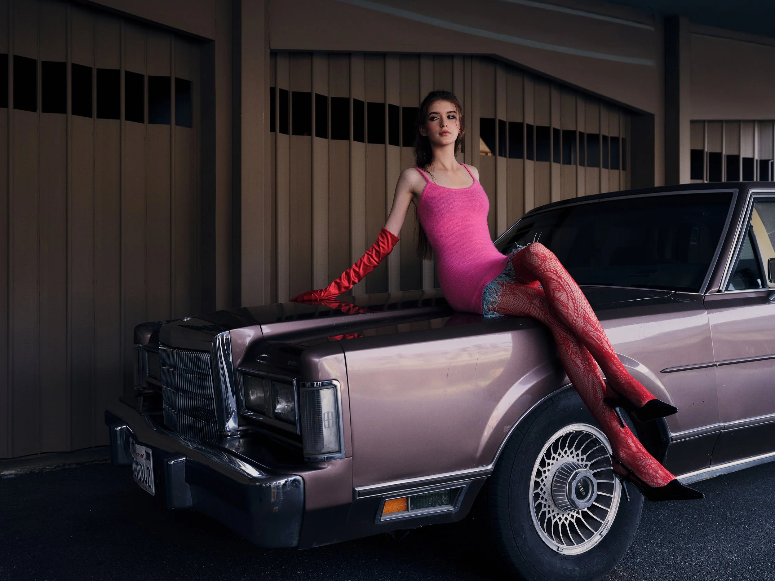 A young woman in a pink dress and red stockings sitting on the hood of a vintage pink car, with her left arm resting on the car and her right arm extended along the car's side, in front of a brown building.
