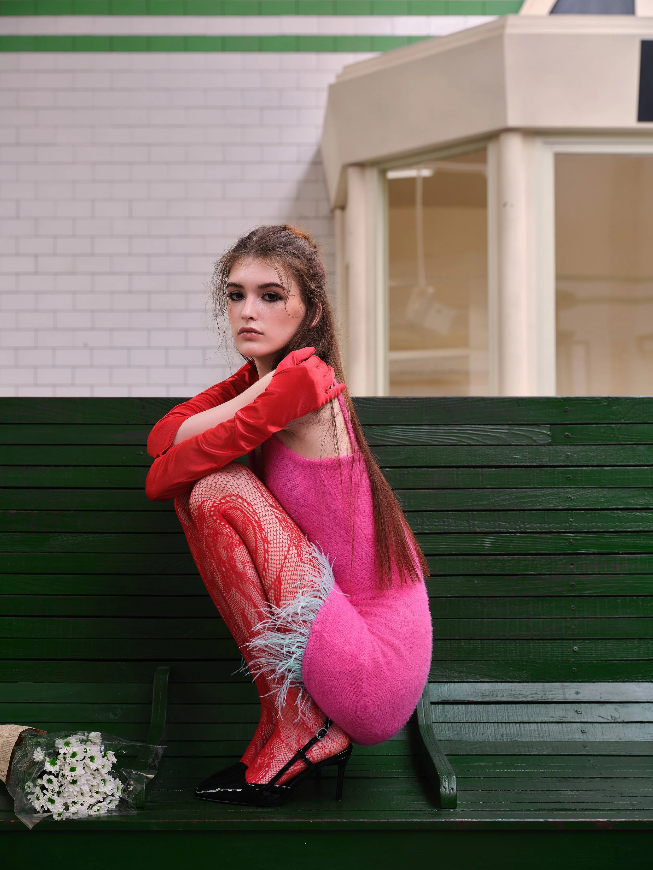 A young woman wearing a pink dress with feathered red fishnet stockings and black high heels, sitting on a green park bench with a bouquet of flowers on the ground next to her.