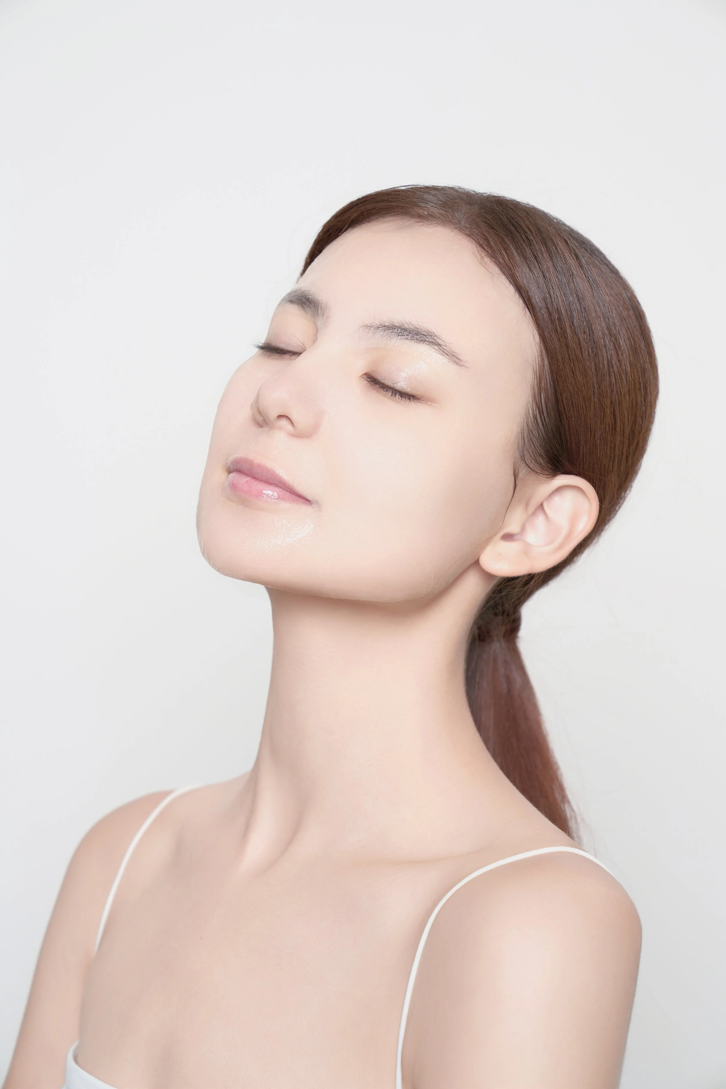 Close-up of a woman with clear skin and brown hair, eyes closed, appearing relaxed against a white background.