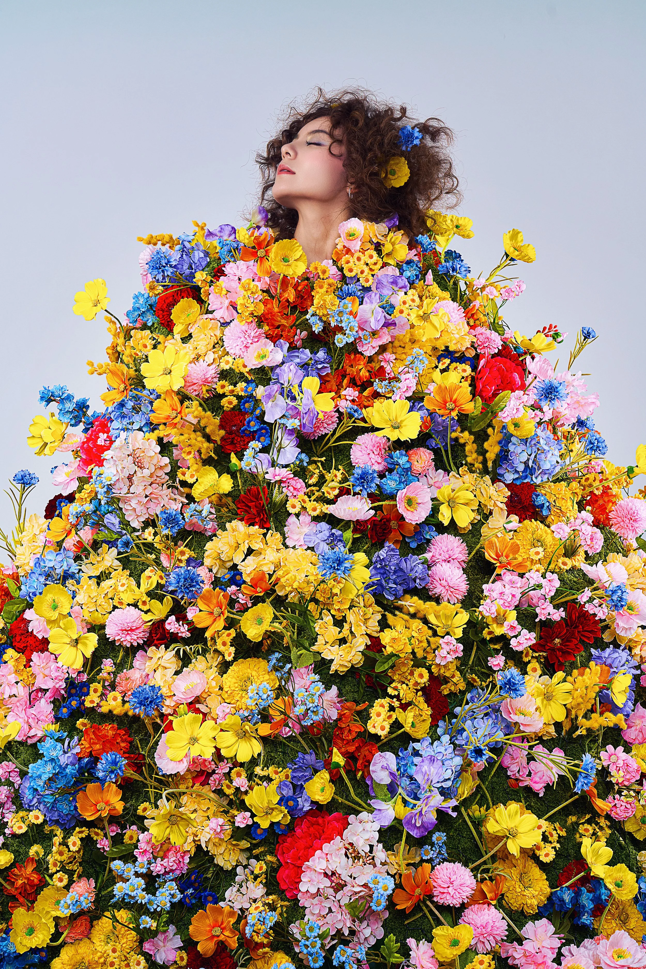 A woman with curly hair and closed eyes surrounded by a large, colorful arrangement of various flowers.