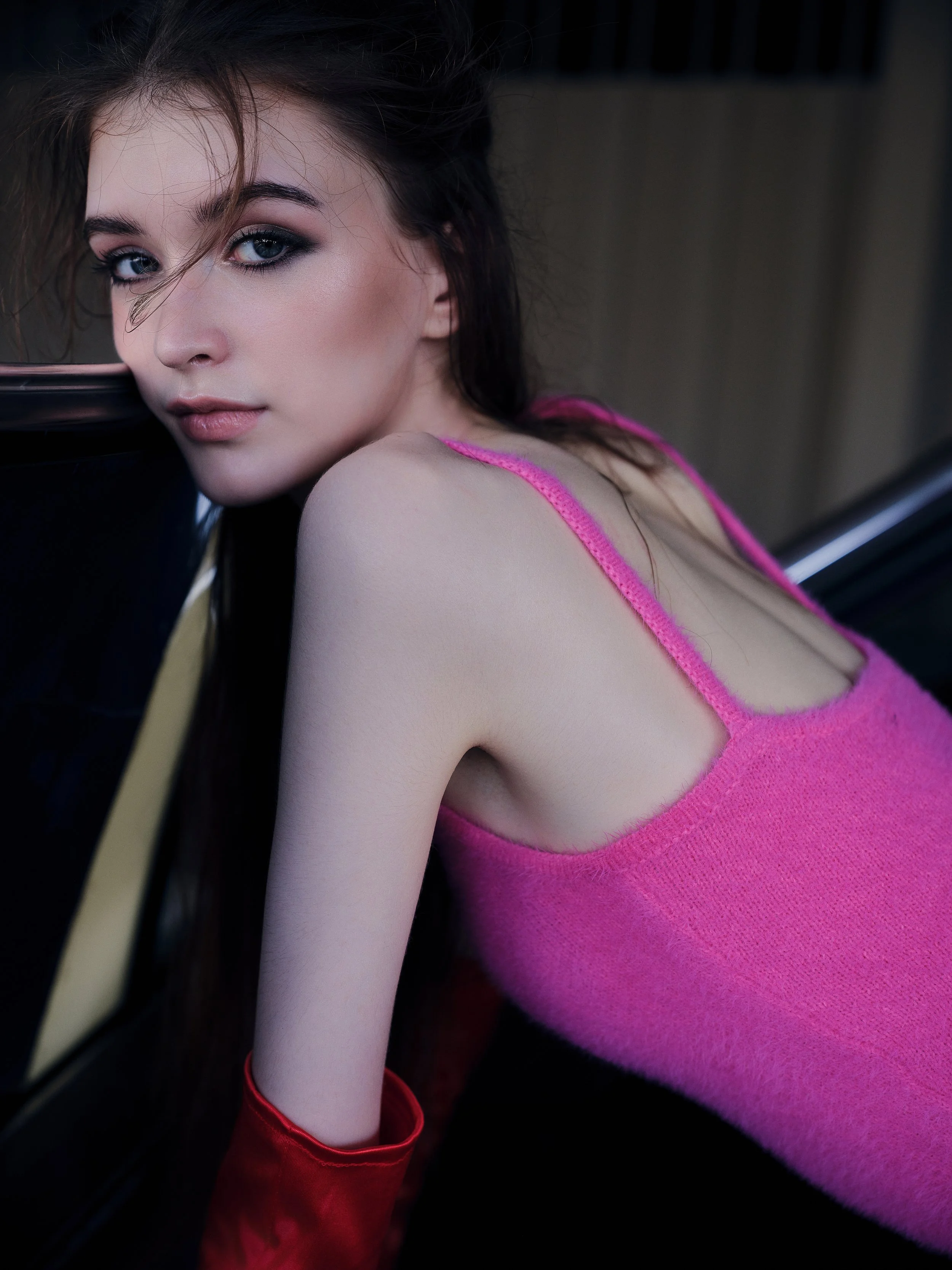 A young woman with long dark hair and blue eyes, wearing a pink tank top and red gloves, leaning her face on a black surface, looking at the camera with a neutral expression.