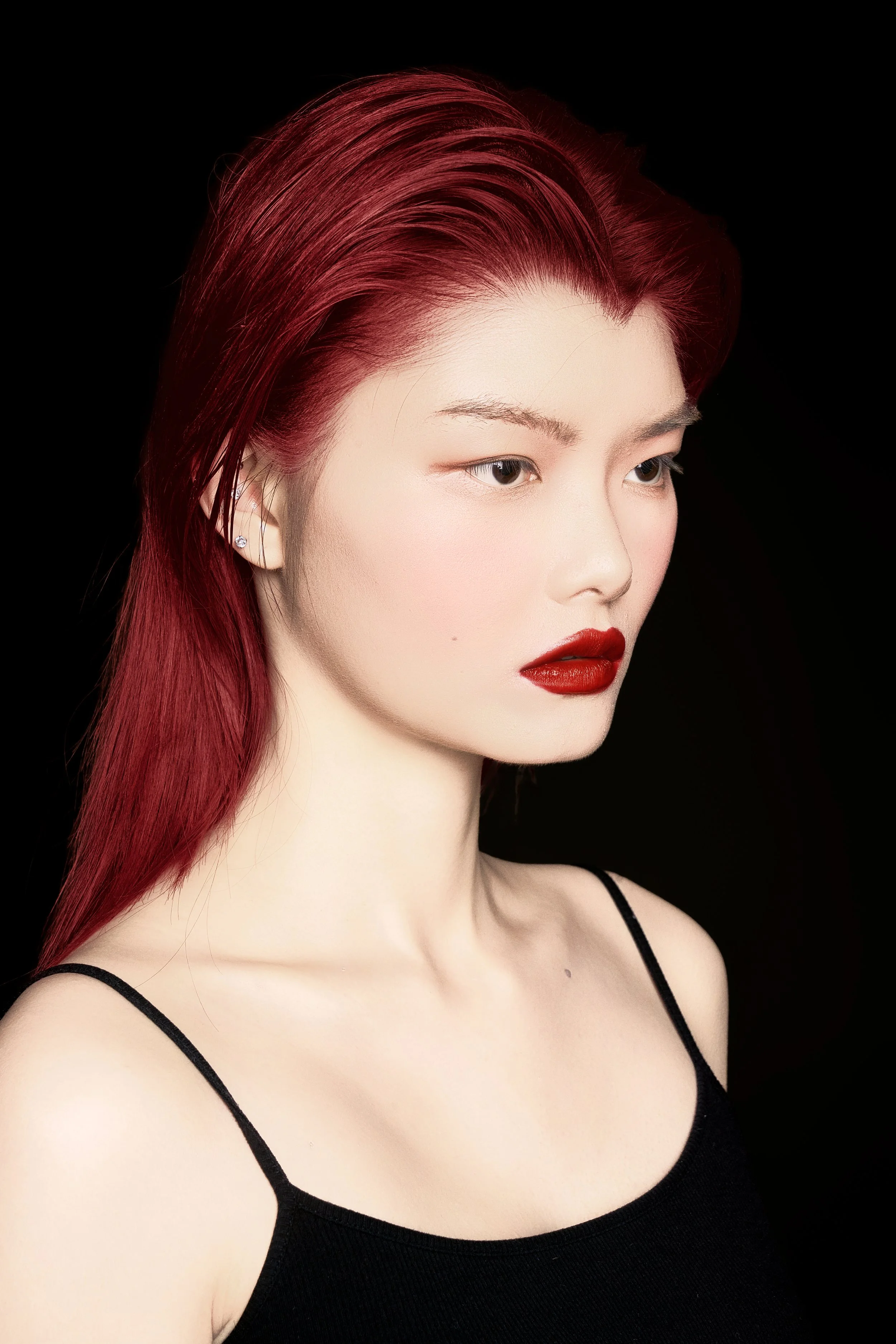 A woman with red hair and makeup, wearing a black spaghetti strap top, against a black background.
