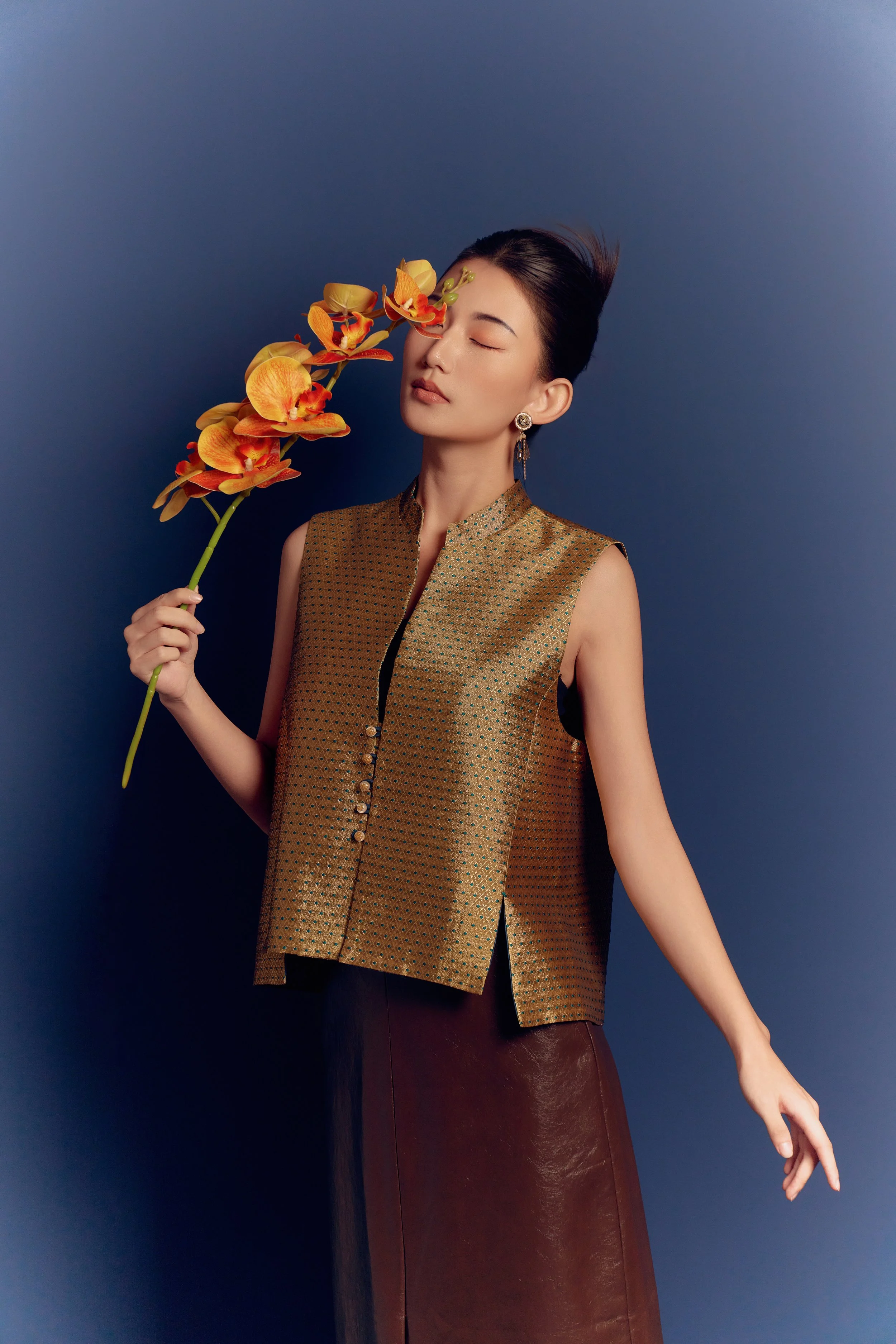 A woman with styled dark hair and earrings holds an orange orchid flower close to her face with her eyes closed against a dark blue background, dressed in a sleeveless metallic gold top and a dark brown skirt.