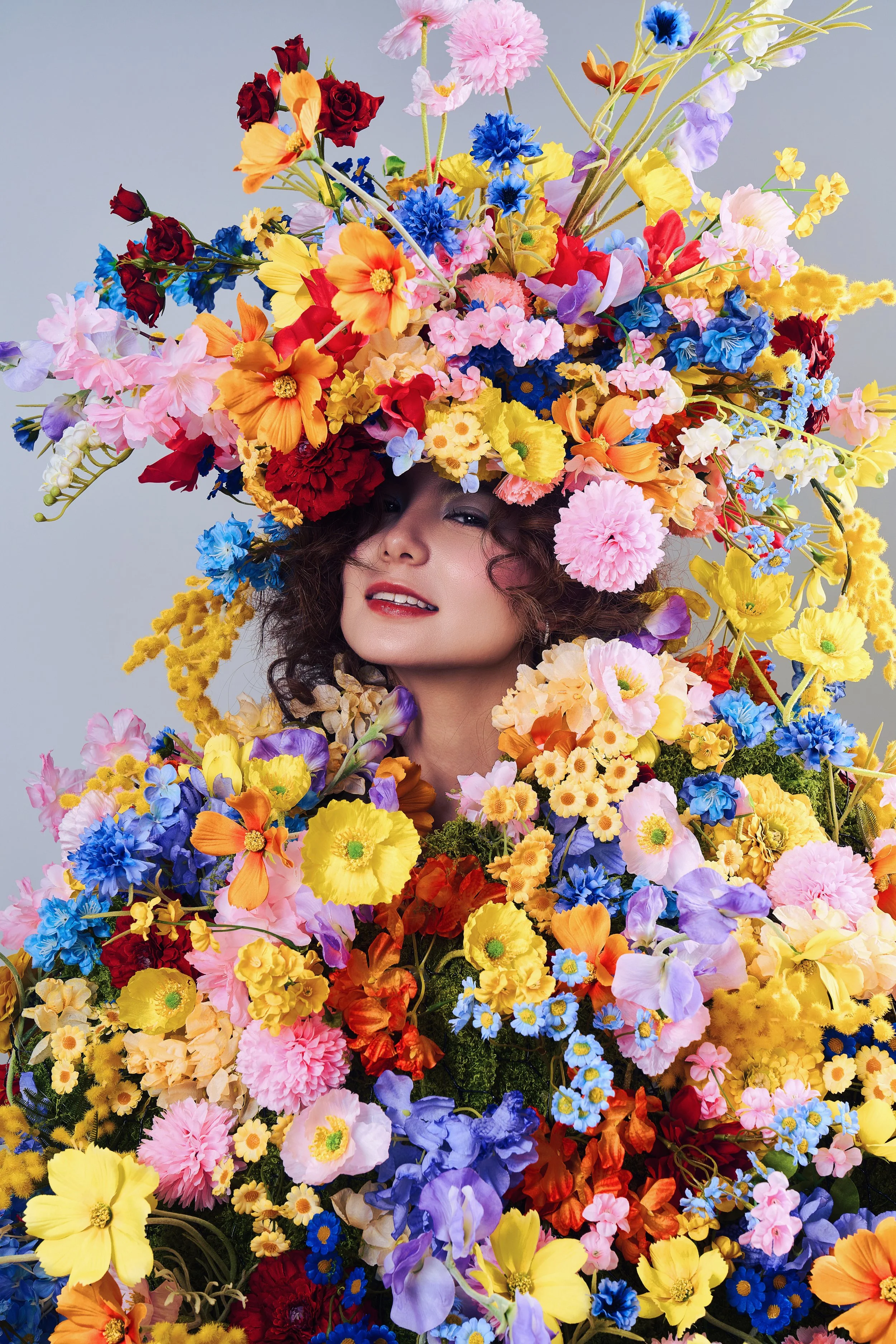 Woman with curly hair surrounded by a large, colorful arrangement of assorted flowers forming a headdress and outfit, against a light background.