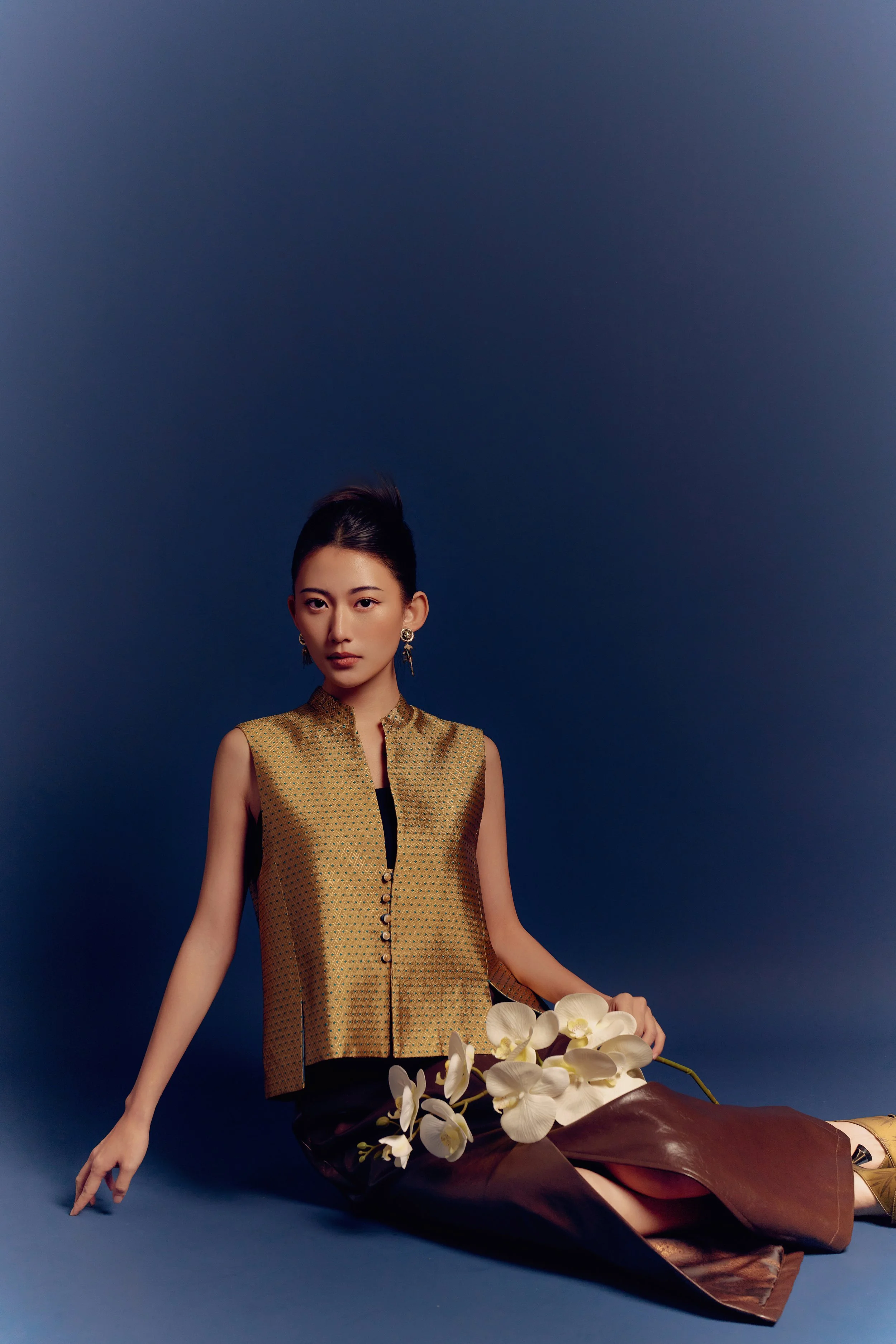 A woman sitting against a plain dark blue background, holding white orchids, wearing a sleeveless gold and black patterned top and dark brown pants, with earrings and styled hair.