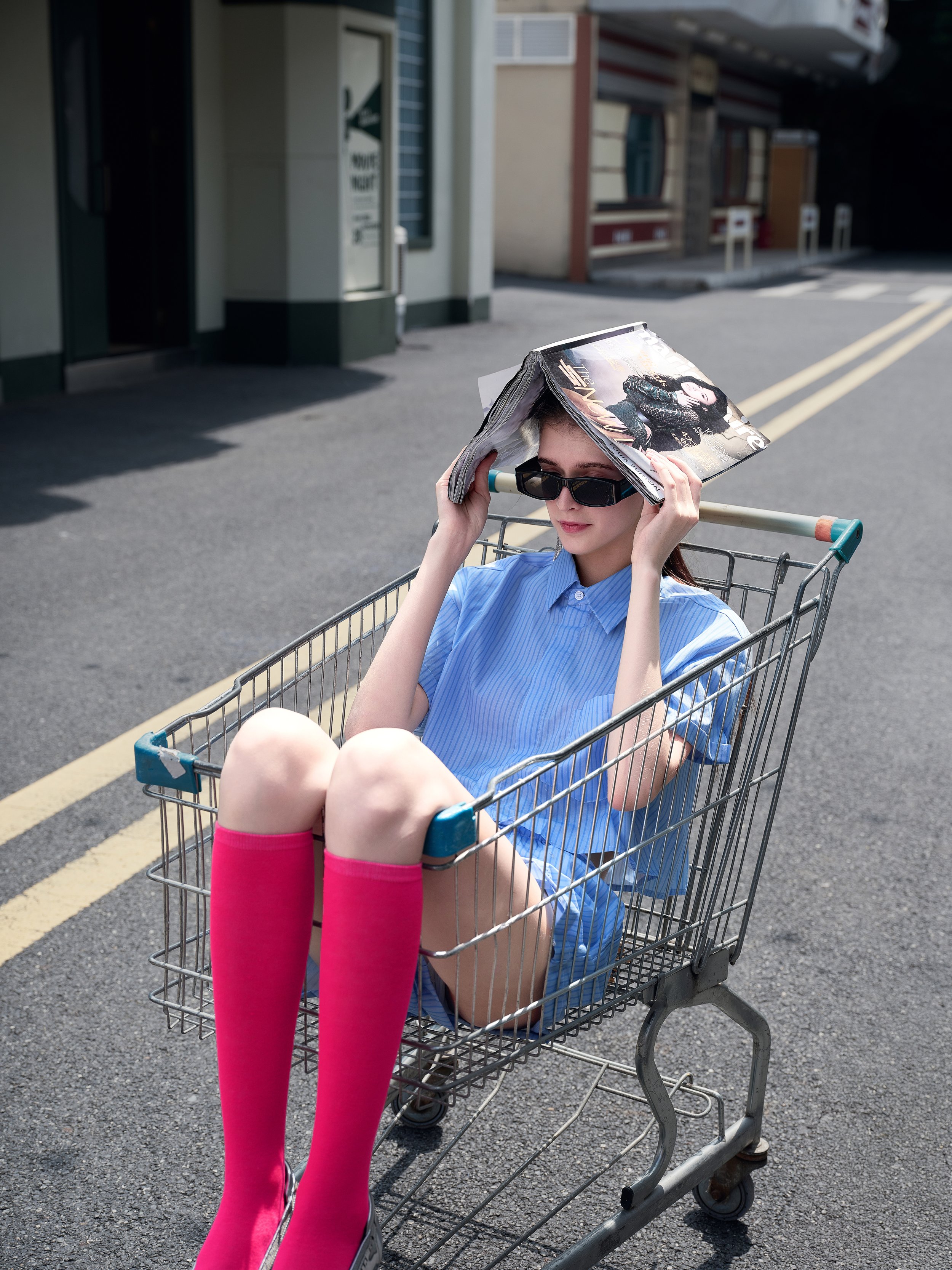 Young woman sitting in a shopping cart on an empty street, wearing sunglasses, a blue striped shirt, pink knee-high socks, and holding a magazine on her head.