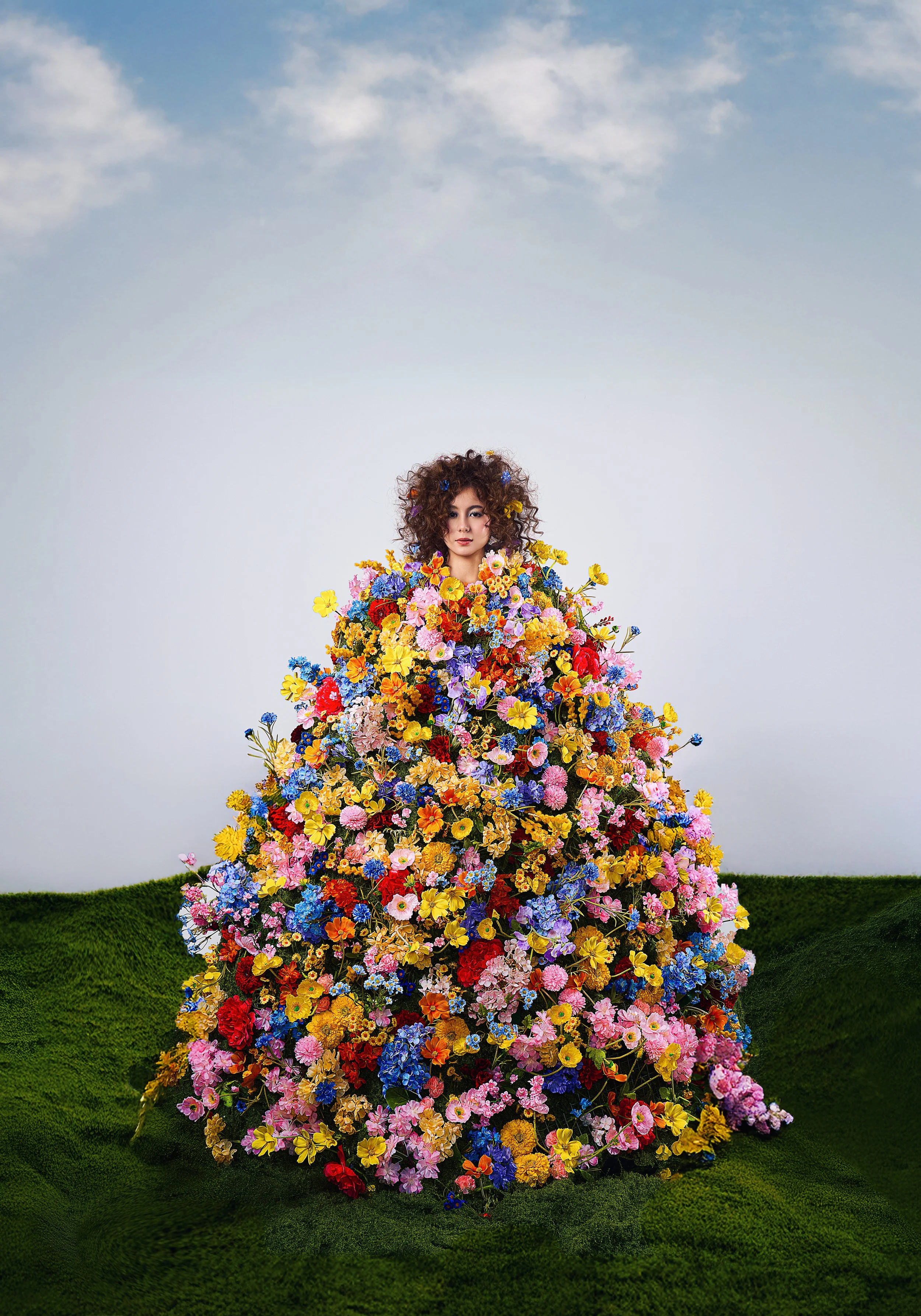 Woman standing behind a dress made of various colorful flowers, on a grassy green surface with a cloudy sky background.