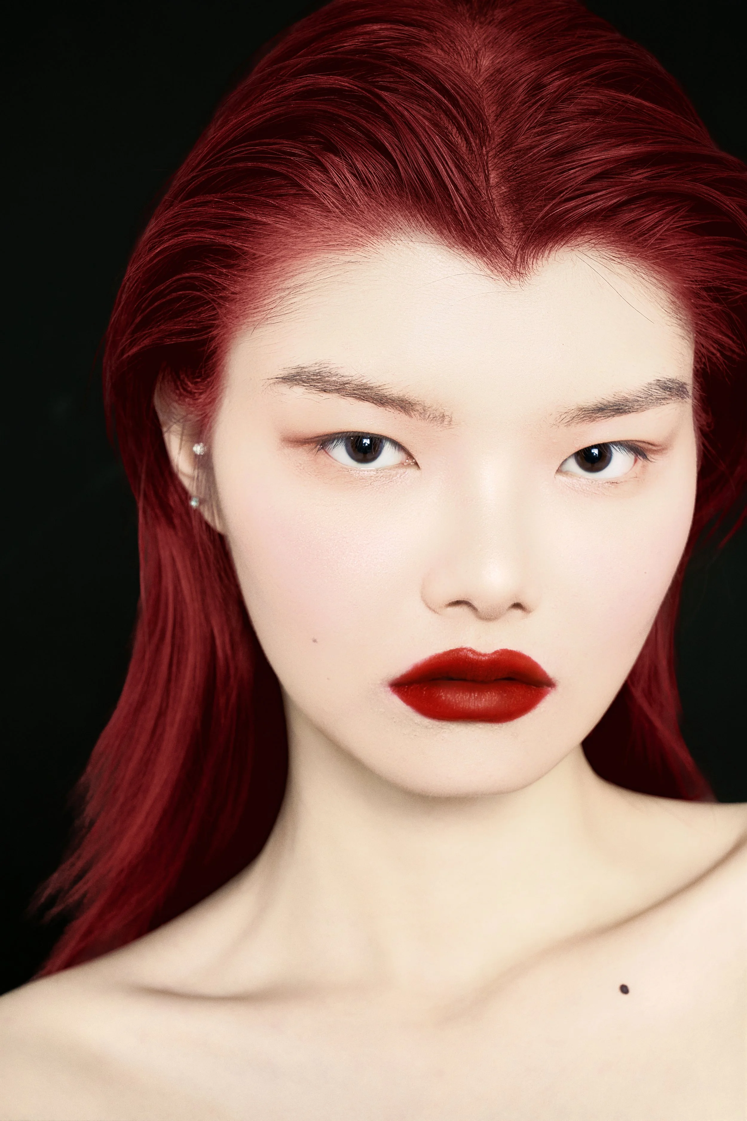 A woman with red hair and bold red lipstick, wearing earrings, against a black background.