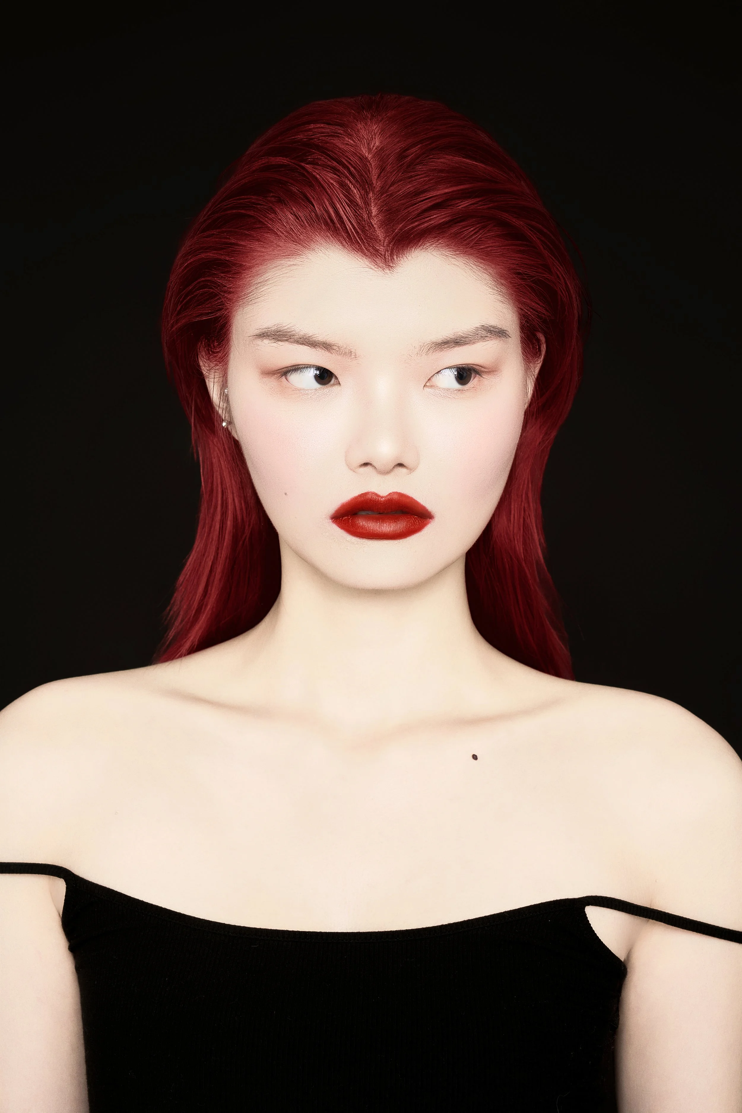 Portrait of a woman with red hair, red lipstick, and a black off-the-shoulder top against a black background.